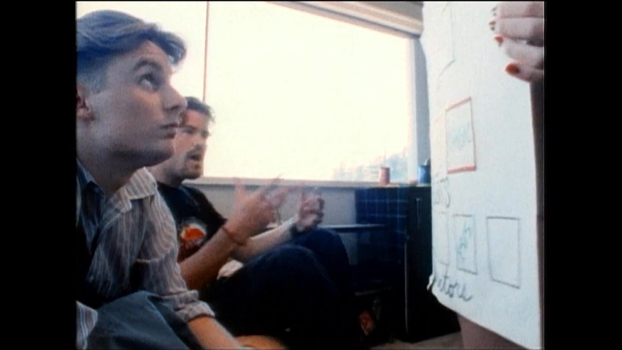 Two individuals engage in a strategic planning session while reviewing names and organizational structure on a display board. The pair discusses project details and professional alignment in a relaxed office environment.
