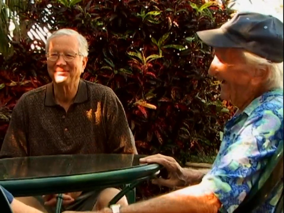 A group of older adults engages in a lively, warm conversation in an outdoor setting. The individuals share stories and laughter while seated at tables in a relaxed, garden-like environment.