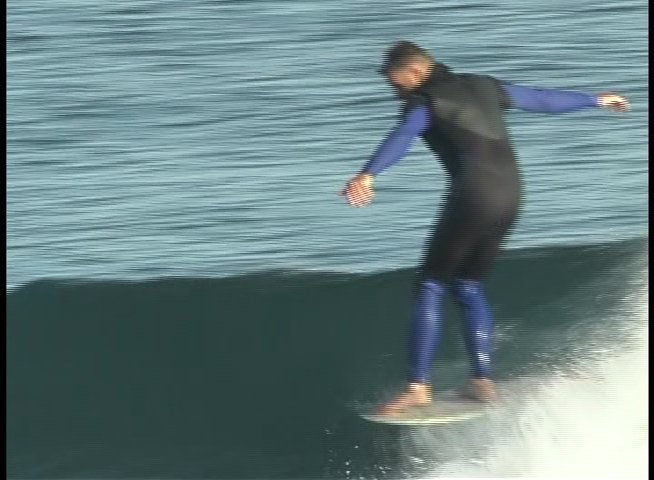 A surfer maneuvers along a breaking wave while wearing a wetsuit, followed by a man speaking during an outdoor interview near a parked truck.