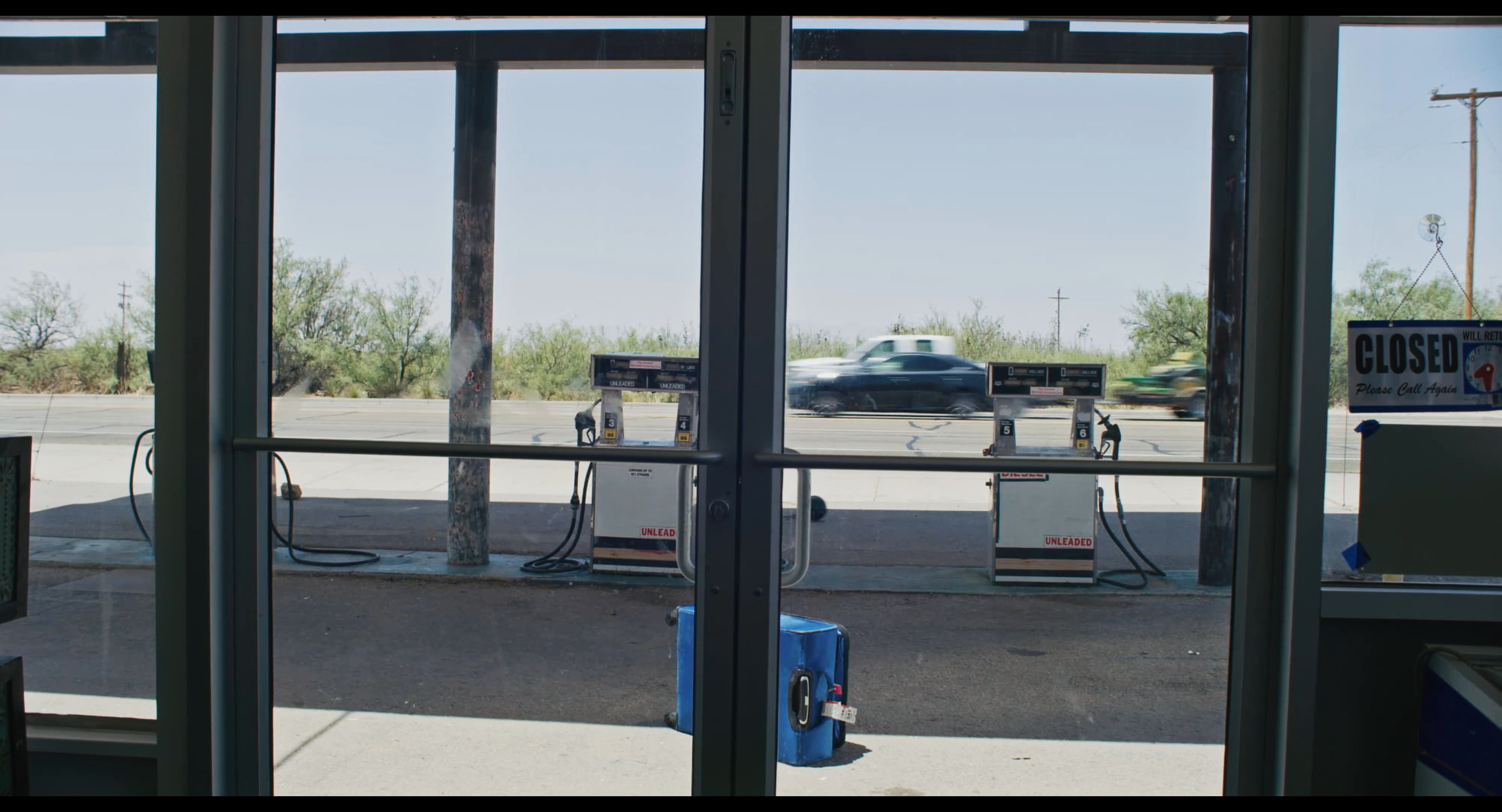 A yellow Mini Cooper pulls into a desert gas station where a passenger exits the vehicle and walks across the pavement. The scene captures a quiet moment of transit in a remote, arid environment.