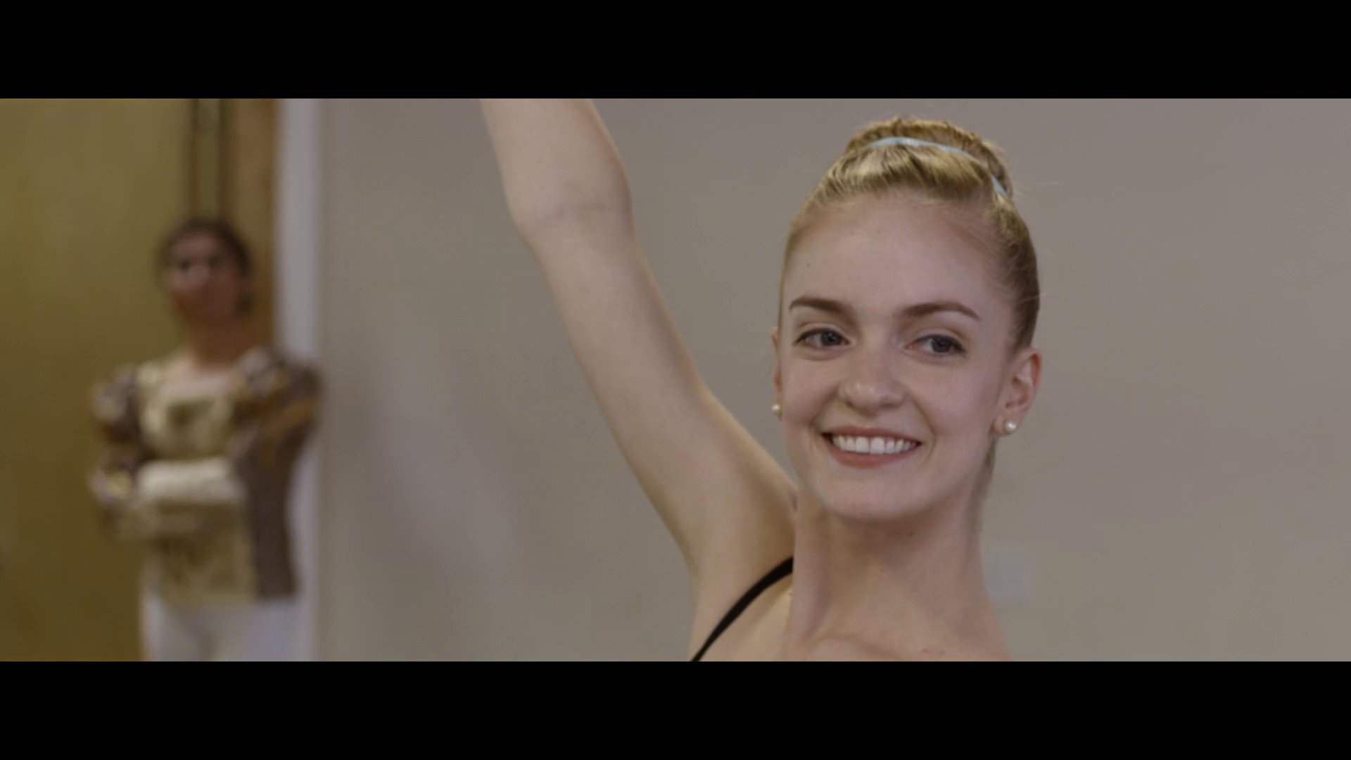 A ballet dancer performs precise movements in a studio setting under the guidance of an instructor. The session transitions from technical practice to a focused interaction between the mentor and the artist.