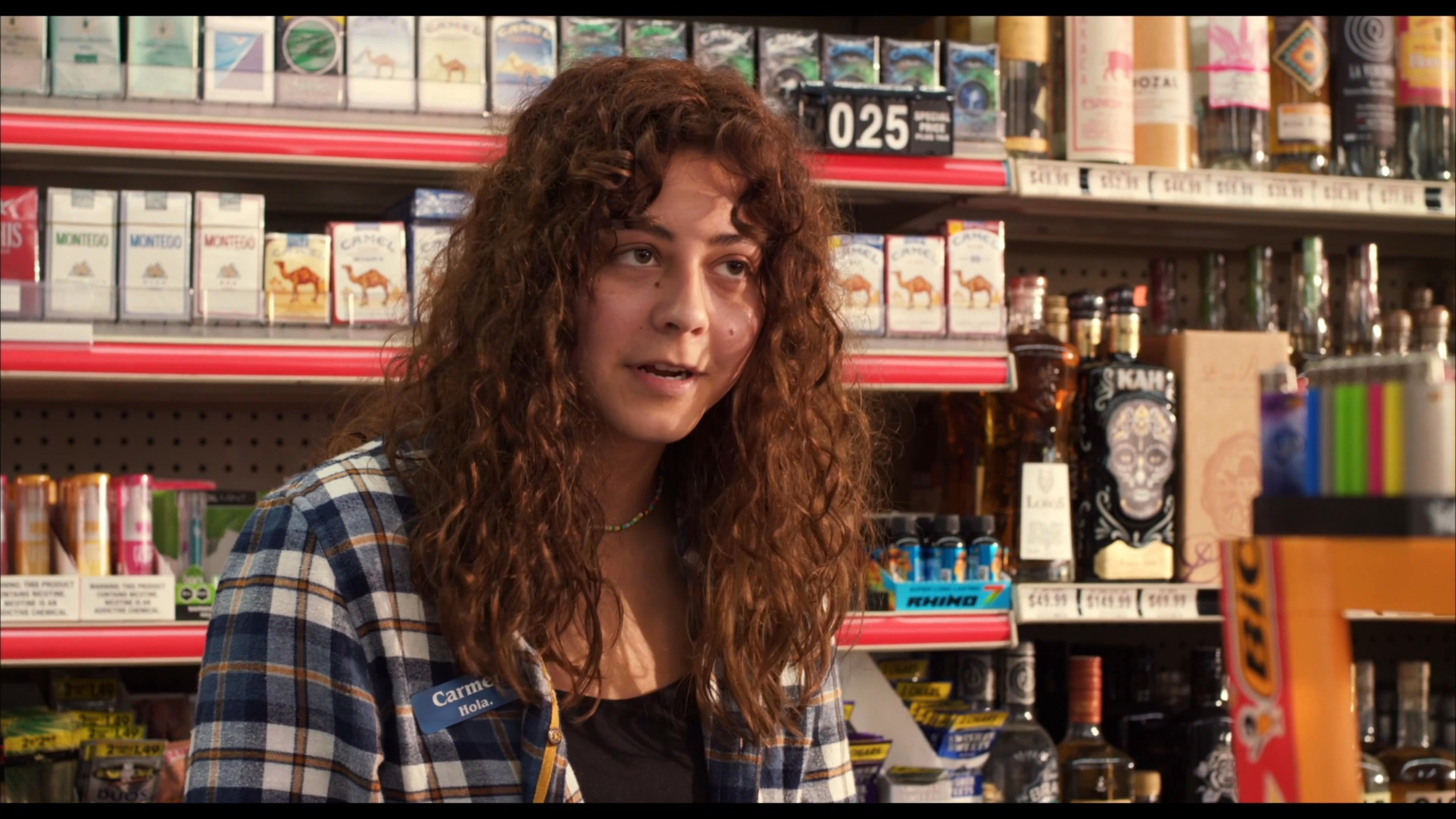A man engages in a dialogue with a convenience store clerk who is browsing a comic book behind the counter. The two maintain a casual exchange within the retail environment.