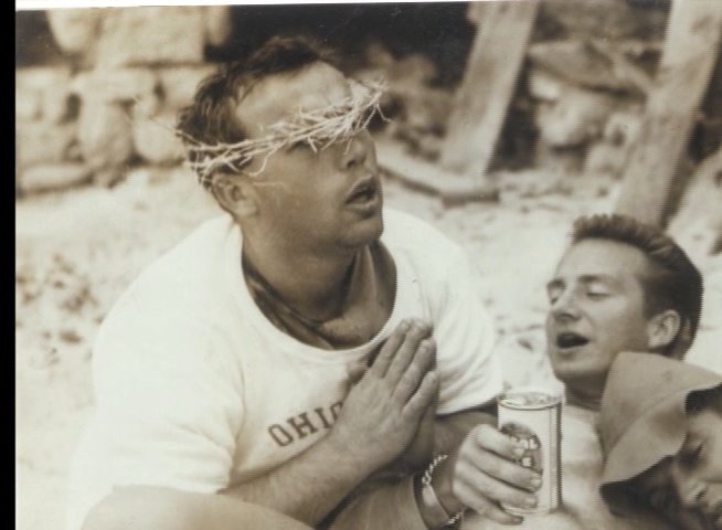 A man wearing a crown of thorns sits with friends in a casual, sunlit outdoor setting. The scene transitions to a candid group of surfers sharing stories and camaraderie.
