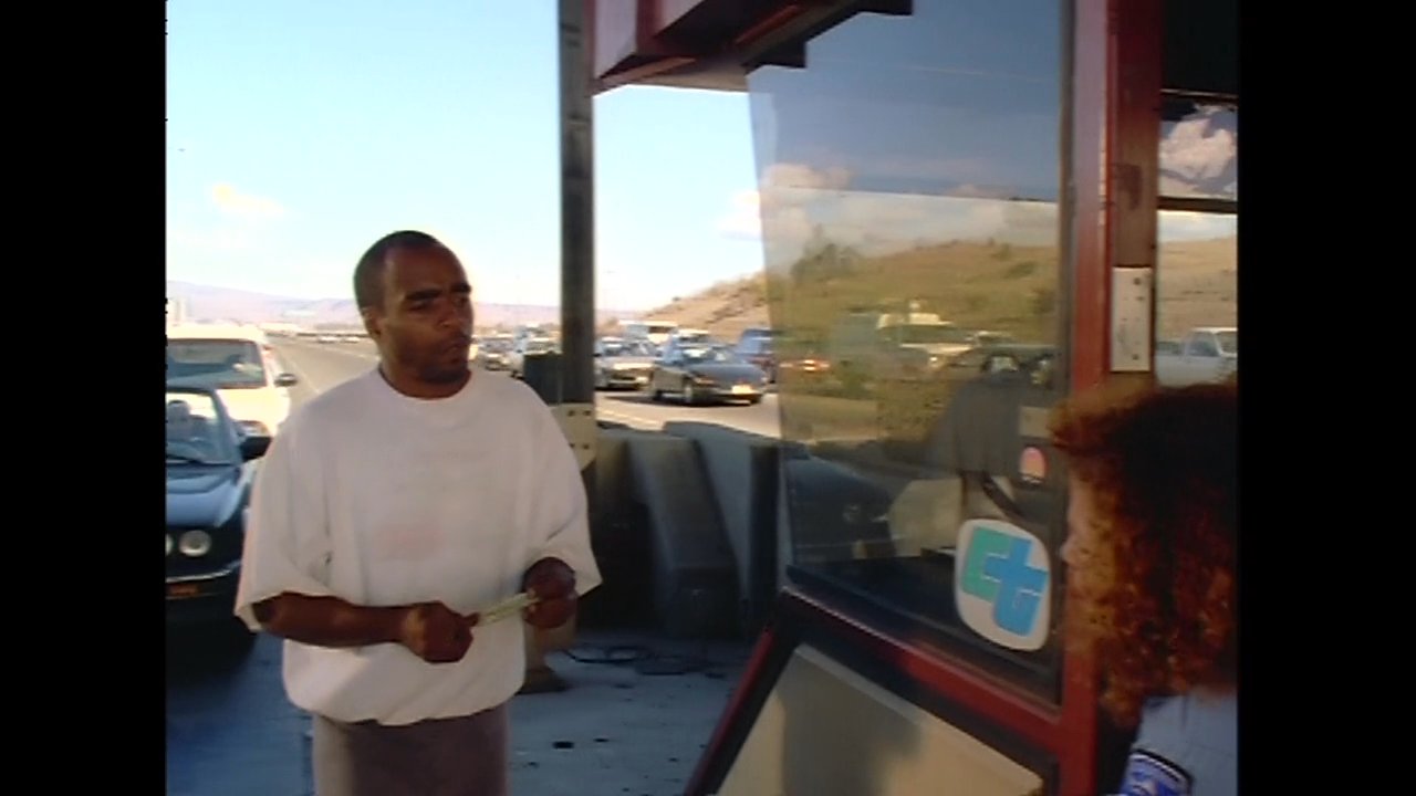 A driver approaches a toll booth and engages in a brief interaction with the attendant to facilitate payment. The exchange occurs on a busy highway under clear daytime conditions.