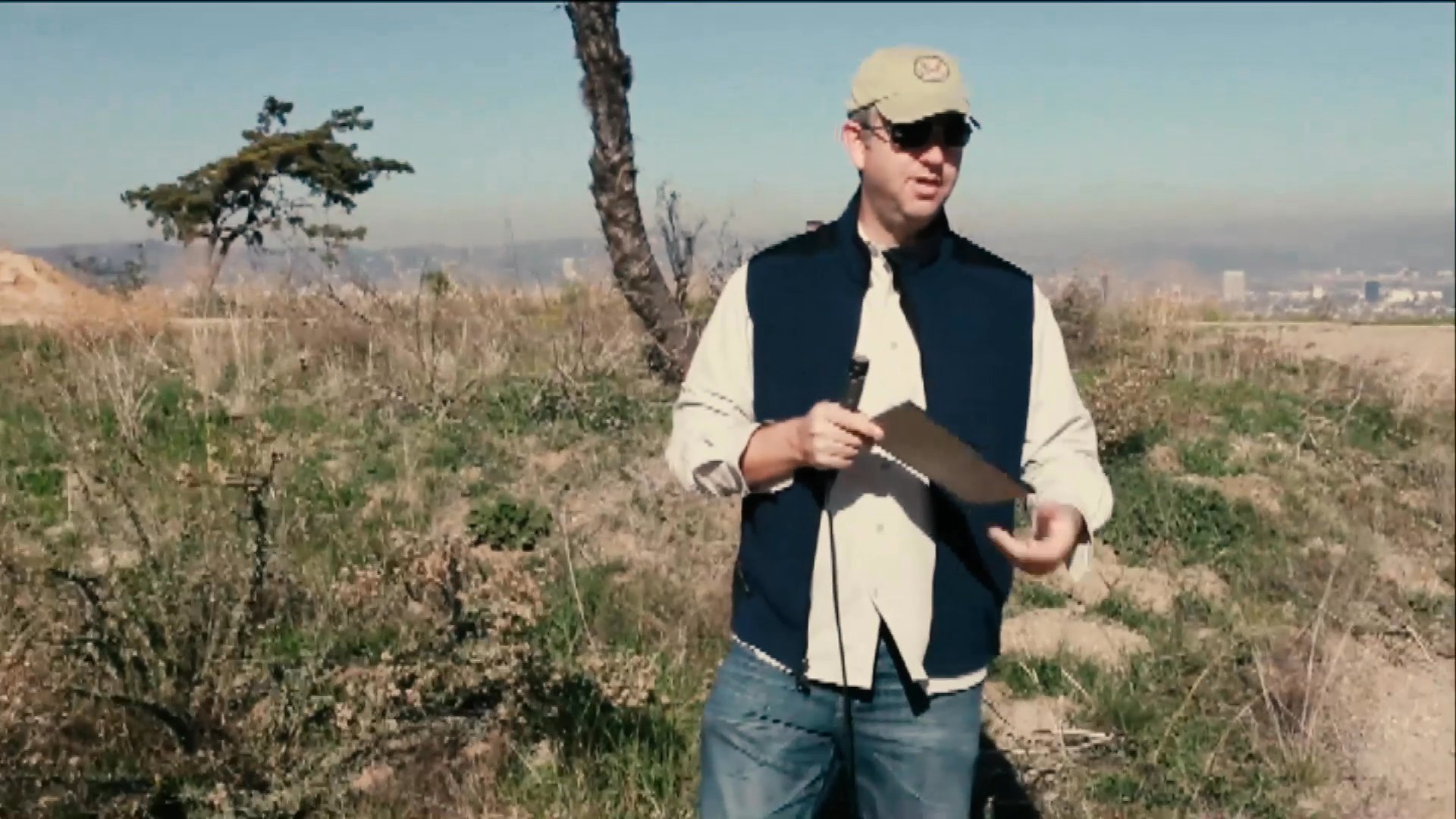 A production crew prepares for a shoot in a scenic outdoor location using a clapperboard to initiate a sequence. The host stands in an open field, coordinating with a technician while reviewing script materials.