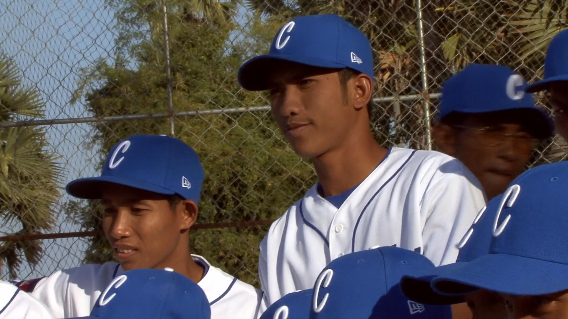 Members of the Cambodia national baseball team gather in their uniforms for a formal team event, interacting with coaches and officials on an outdoor field.