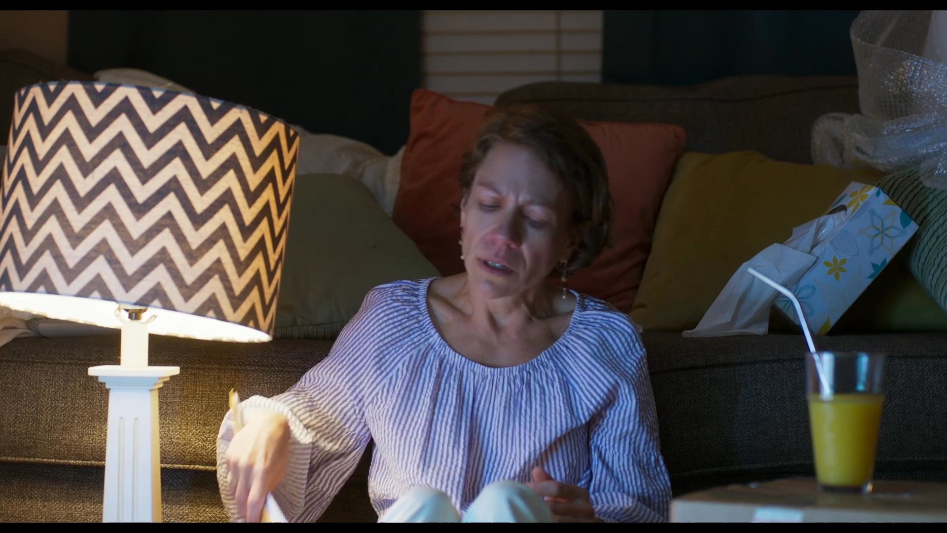 A woman experiences a moment of profound sadness while sitting alone in a dimly lit living room, holding documents. The intimate setting highlights her somber mood during a quiet evening.