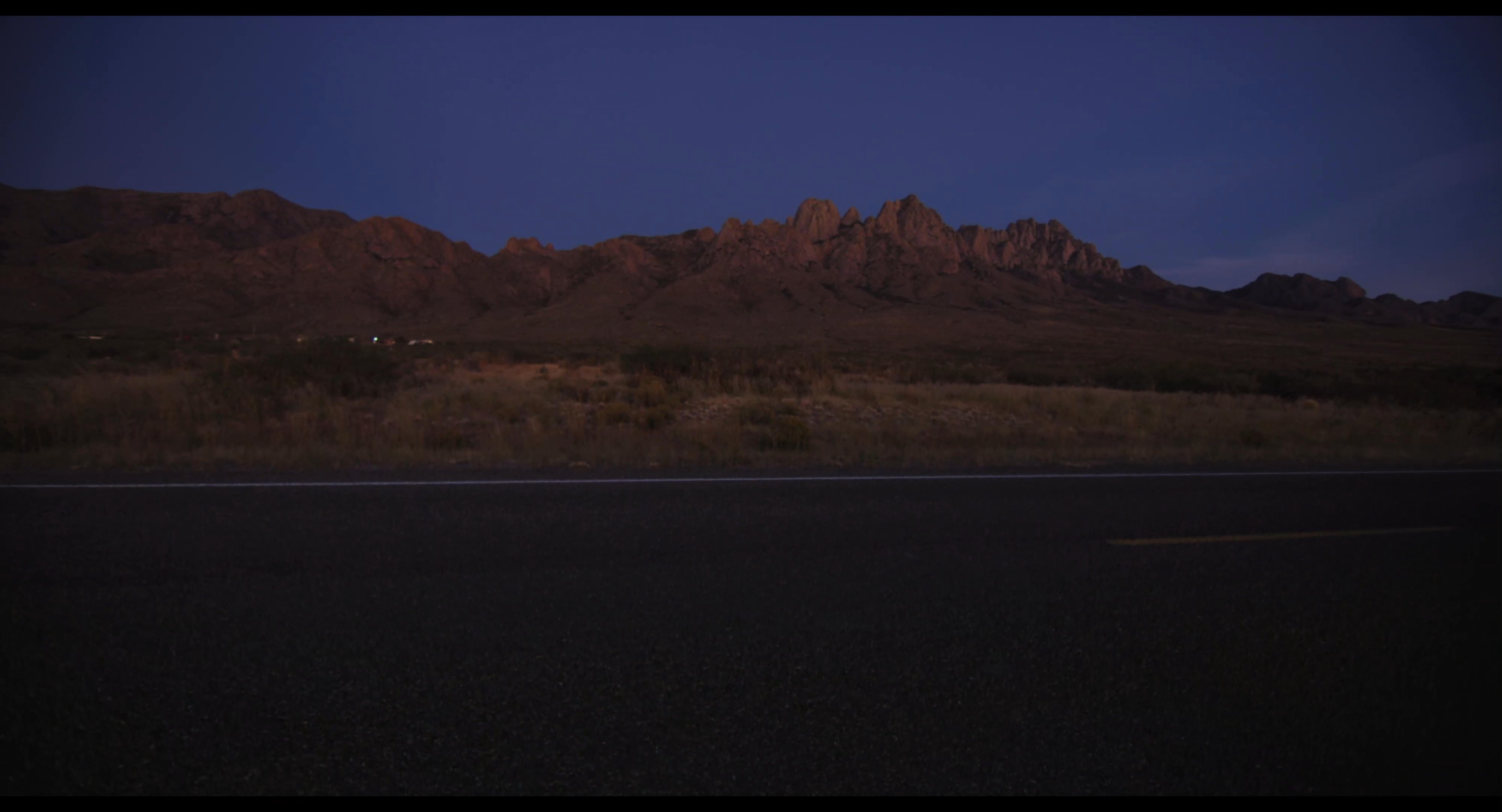 Daylight fades over a vast desert landscape as twilight gives way to the night. The sequence concludes with a quiet view of a lit gas station under the evening sky.