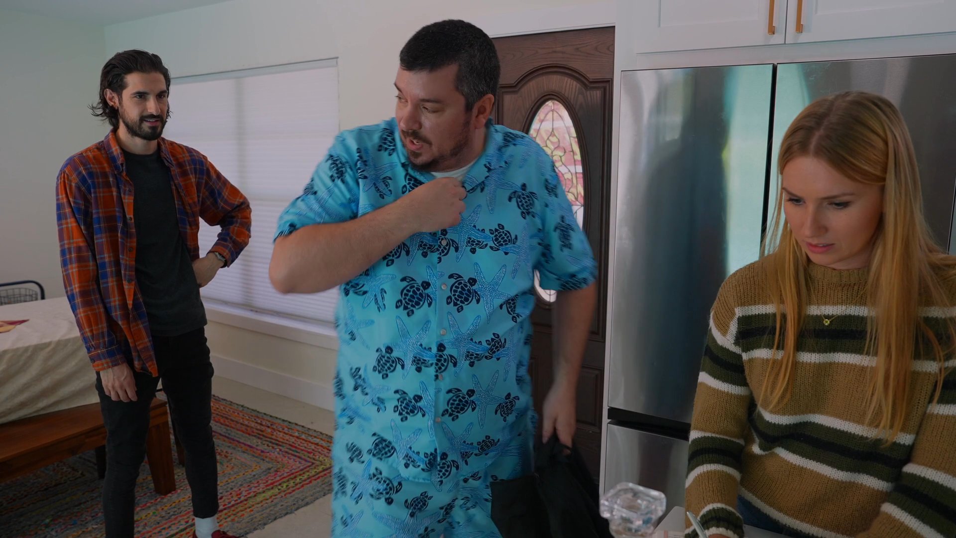 A man in a floral outfit unpacks items from a bag and pours a drink while a woman looks on in a residential interior. The scene continues with the man interacting with others present in the home environment.