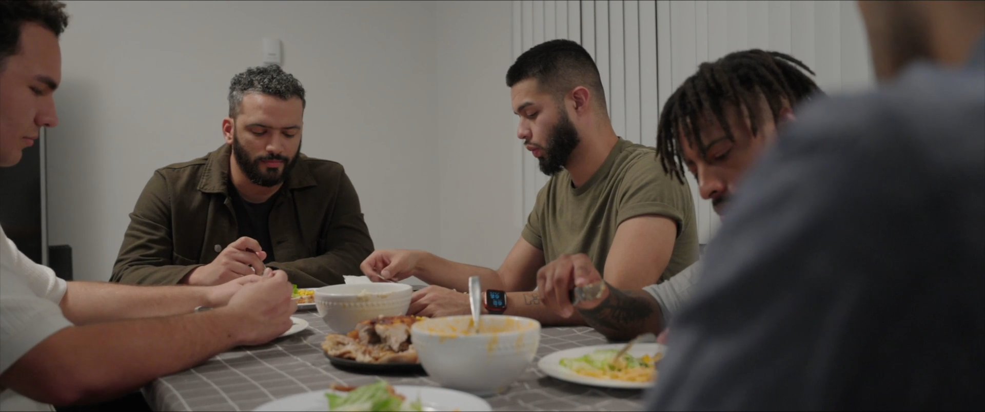A man shares a message on his smartphone with friends gathered around a dinner table, leading to a group discussion while they eat. The interaction shifts from digital communication to collective social engagement during a meal.