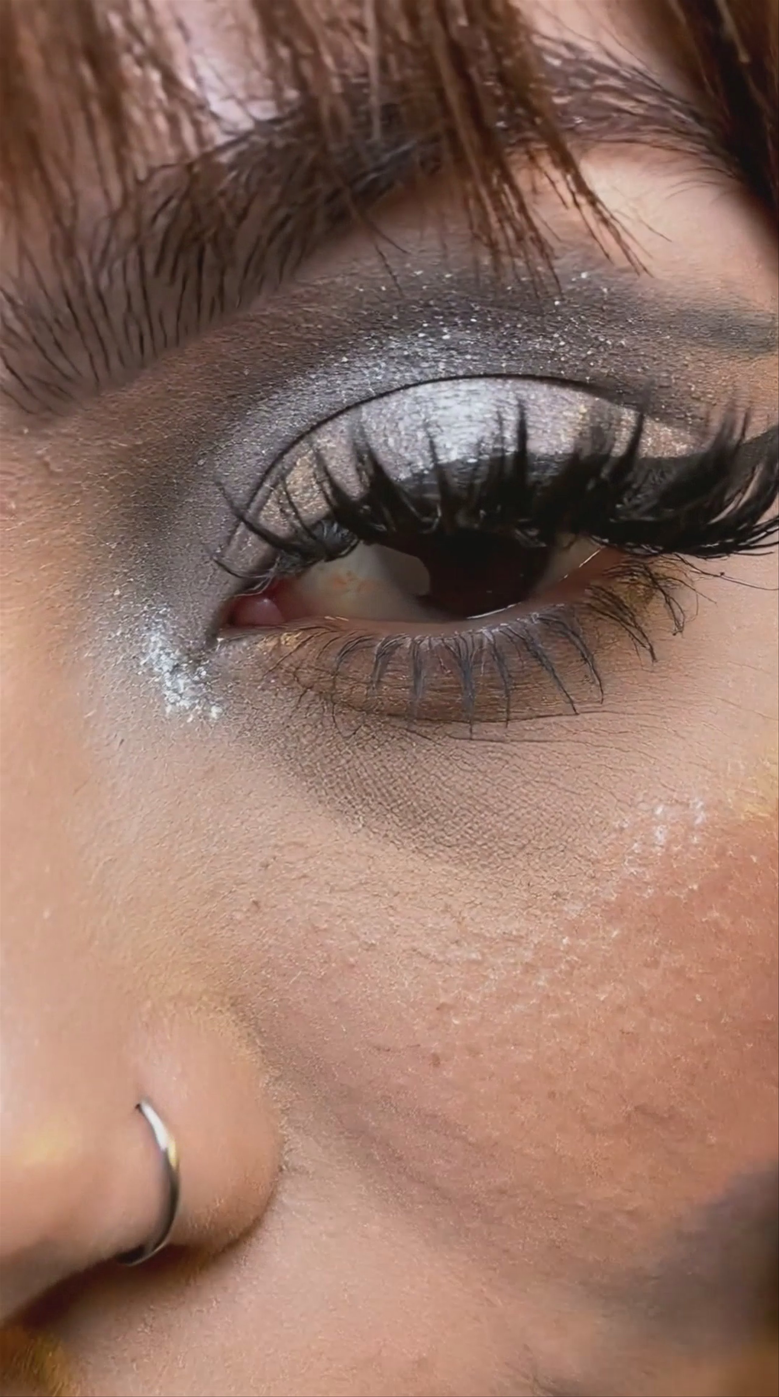A close-up vertical video frame of a young woman with brown hair and bangs, wearing shimmering silver eyeshadow and a silver chainmail halter top. She poses with her hand near her face against a warm yellow backdrop. This 1076x1928 video was generated using the Kling O1 model on Hedra.