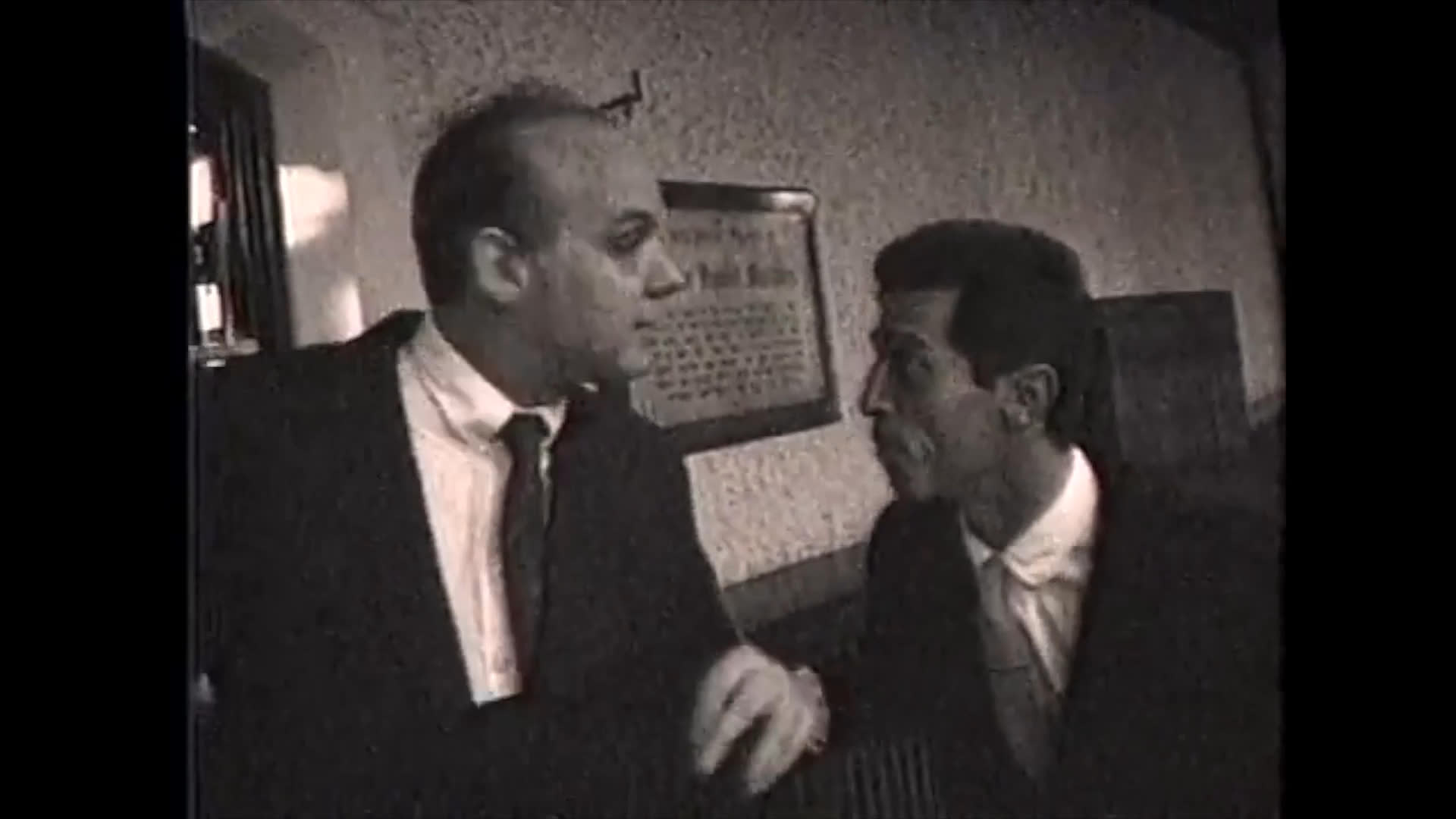 Two men in business attire engage in an intense, conversational exchange within a vintage-style interior setting. The sequence captures their evolving expressions and gestures as they interact in a formal environment.