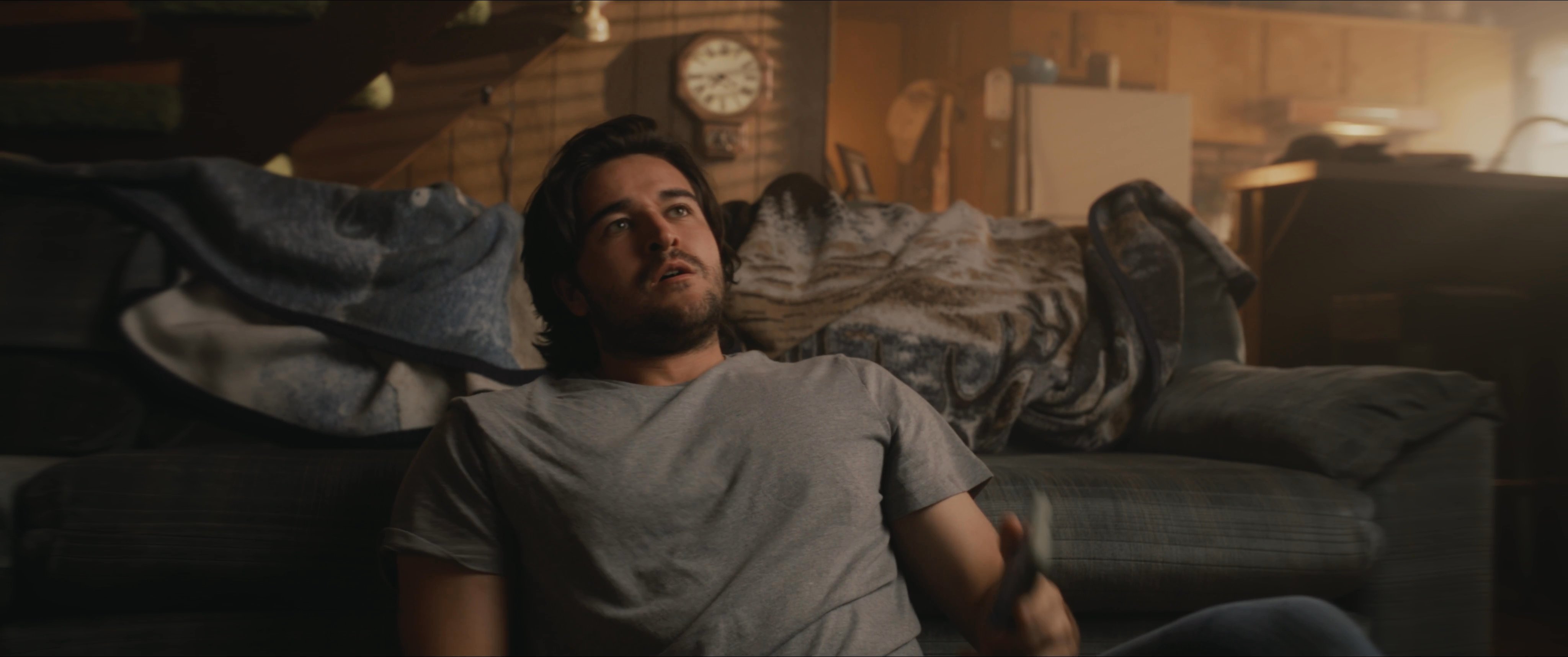 A man resting on the floor in a dim living room answers a phone call from Mark Fischer. He engages in a quiet conversation while reclining against his sofa.
