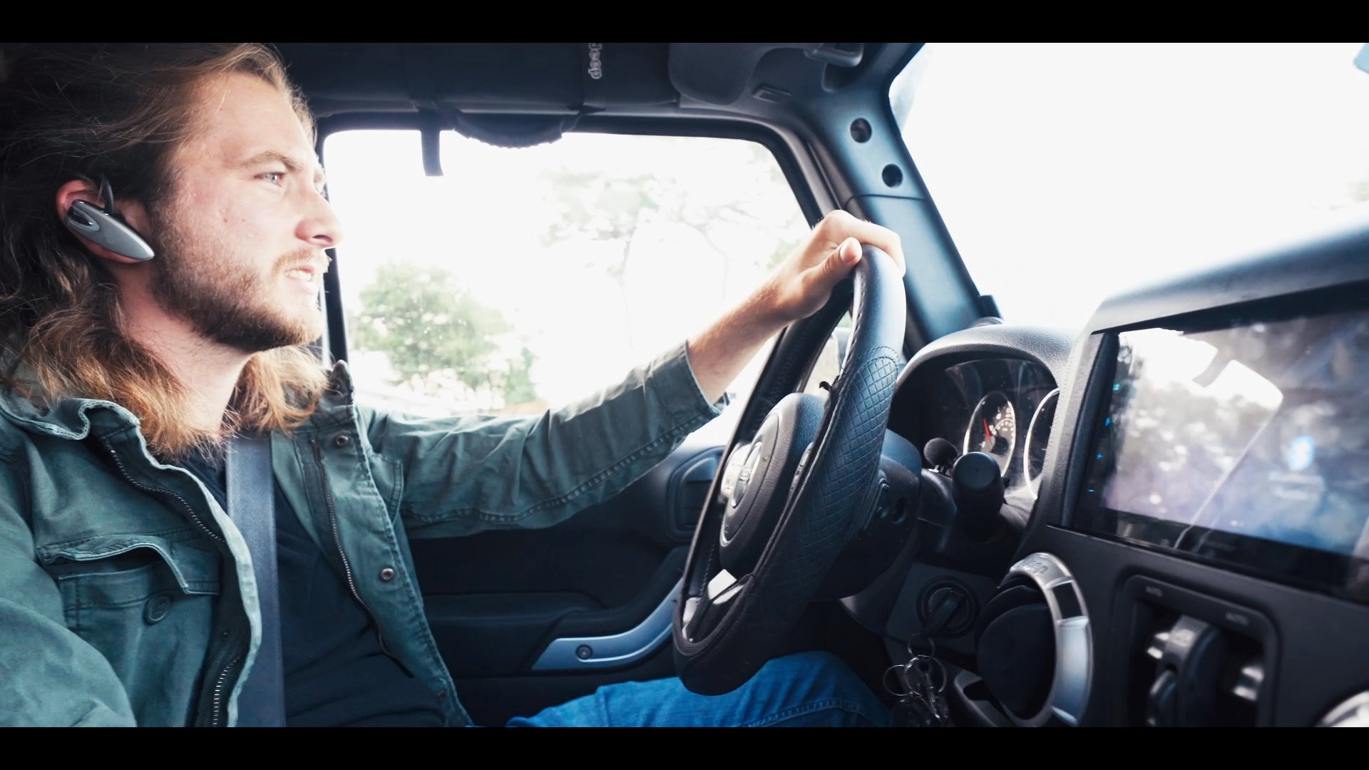 A man with long hair drives a vehicle while engaged in a conversation through a bluetooth headset. He maintains focus on the road and speaks animatedly during his commute.