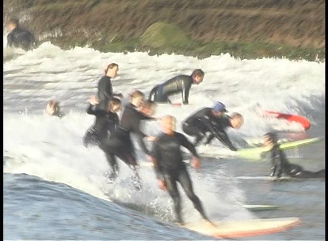 Surfers ride waves in a coastal setting, demonstrating balance and technique while navigating the break. The footage highlights multiple individuals engaging in surfing activities alongside a rocky shoreline.