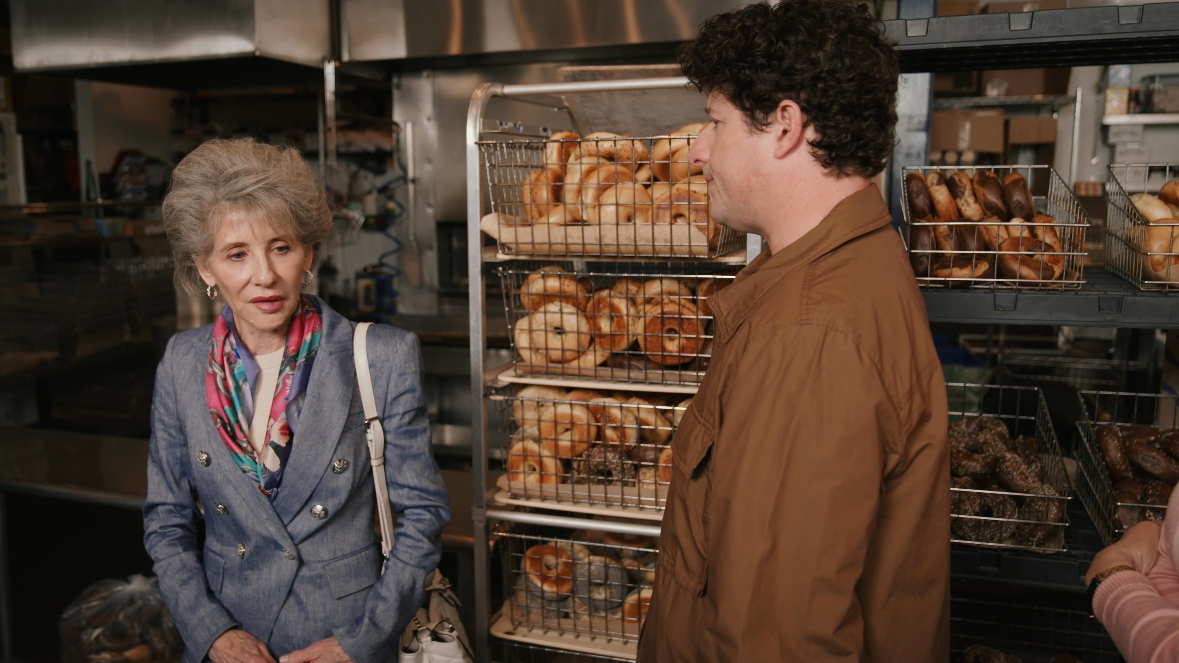 A man engages in a focused conversation with an older woman inside a bakery. The interaction continues as they exchange words in front of stocked shelving.