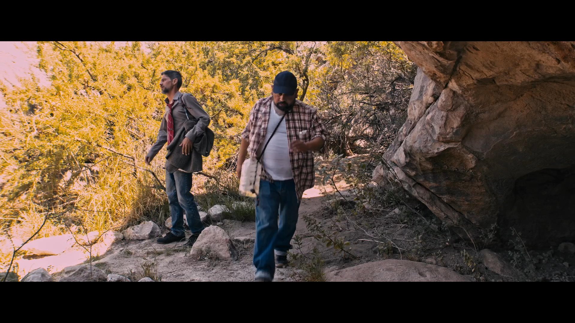 Two individuals hike through a rugged, arid landscape before stopping to rest and drink water near a rock formation.