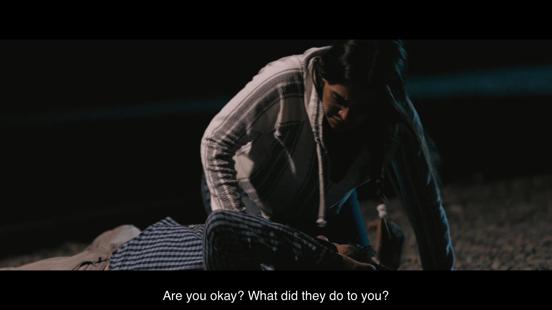 A woman discovers and comforts an injured man lying on the ground at night. The pair exchange tense dialogue regarding a recent encounter before the man attempts to rise.