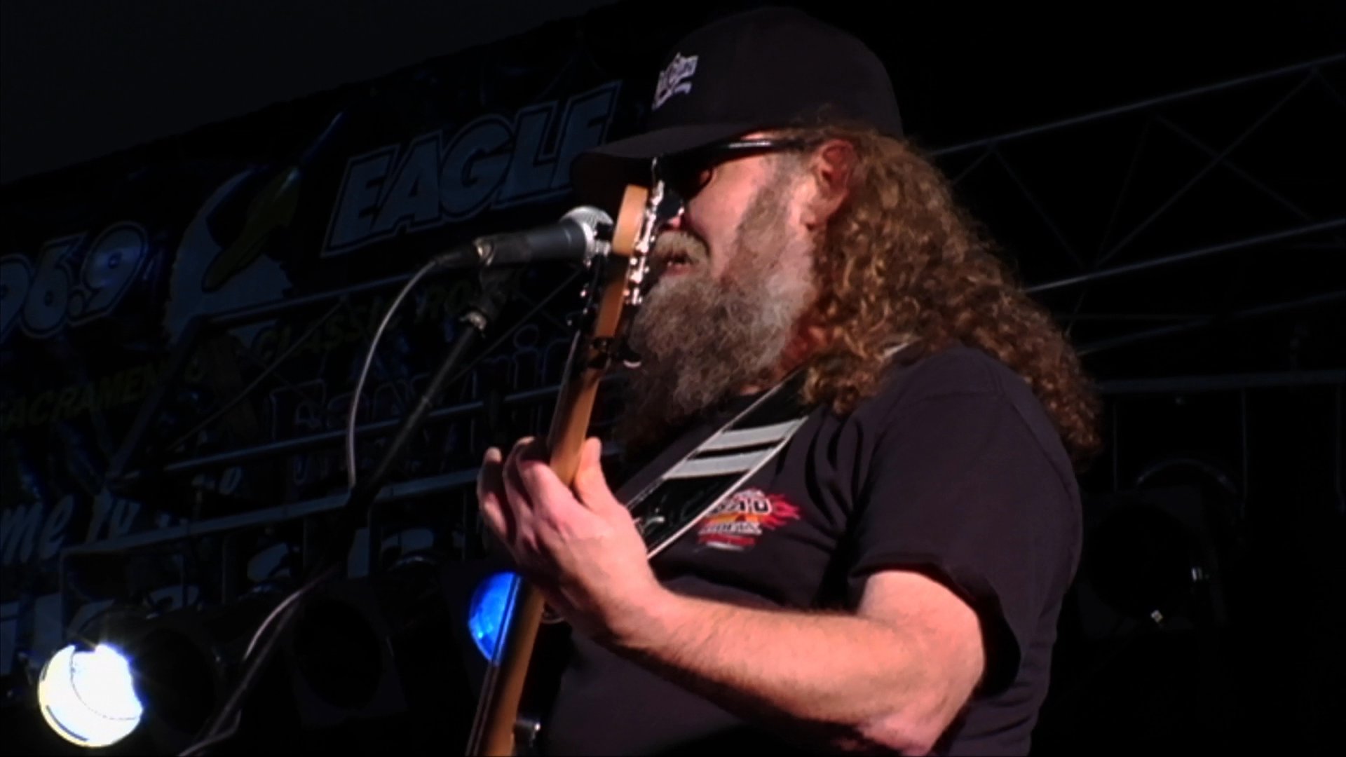 A long-haired, bearded guitarist and a bassist perform on a dimly lit stage. The musicians play their instruments while singing into microphones during a live concert.