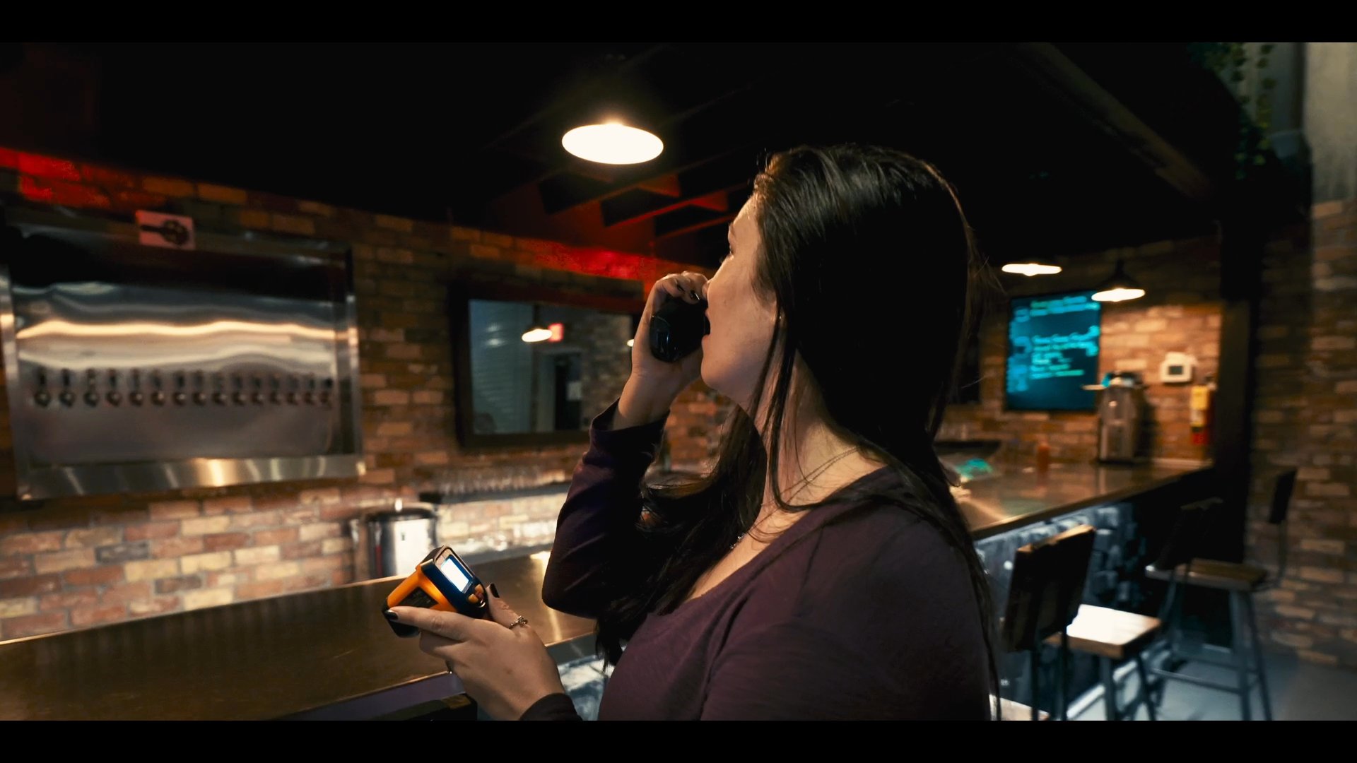 A woman stands in a dimly lit bar interior, holding a two-way radio to her ear while focusing on her communications. She maintains a professional demeanor while coordinating tasks within the establishment.