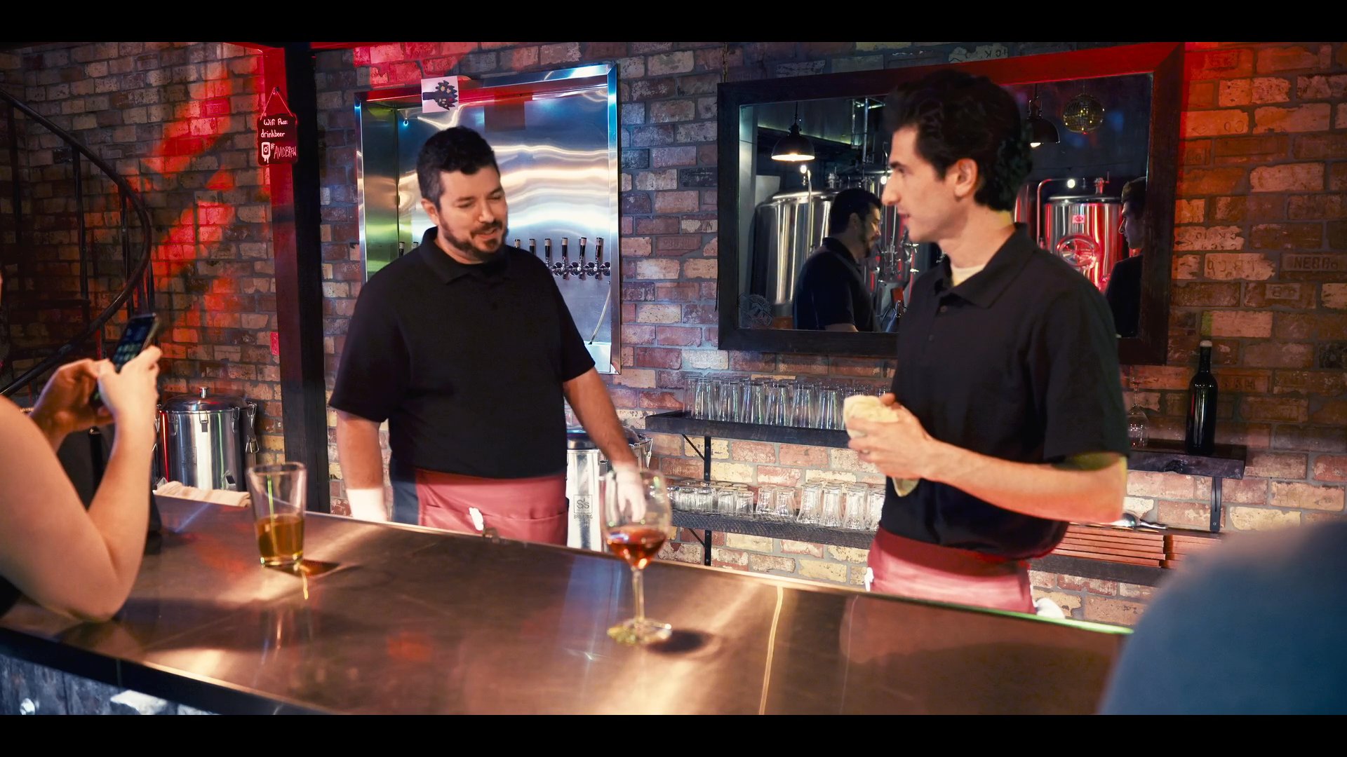 A bartender wipes down a brewery counter and organizes glassware before engaging in a professional conversation with a colleague.