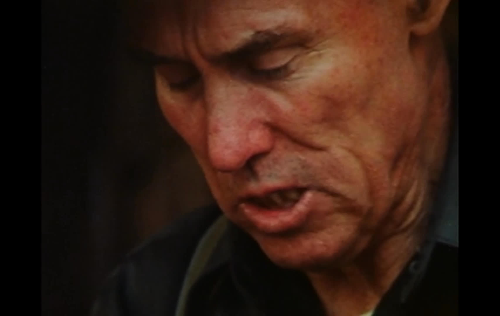 An older man in a cowboy hat sings a country-style song in a dim, rustic workspace. He works with his hands at a bench as the view widens from close-ups to a medium shot.