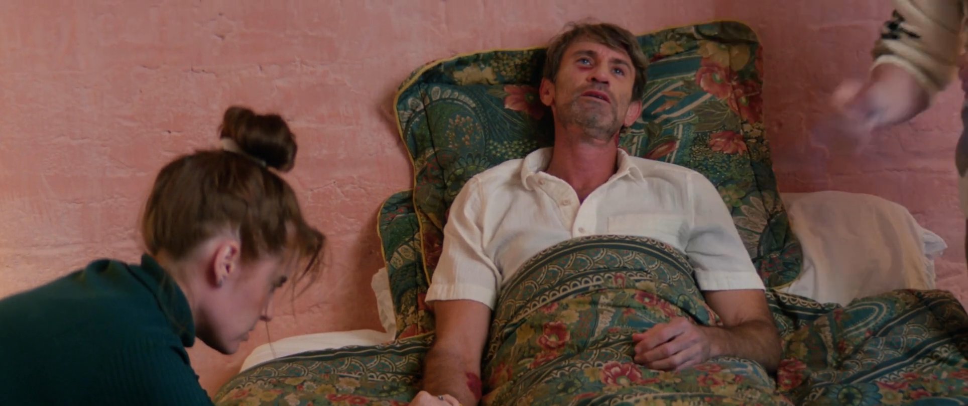 Family members provide comfort and support to a man resting in bed within a home setting. The sequence depicts expressions of care and physical reassurance during an intimate moment of recovery.
