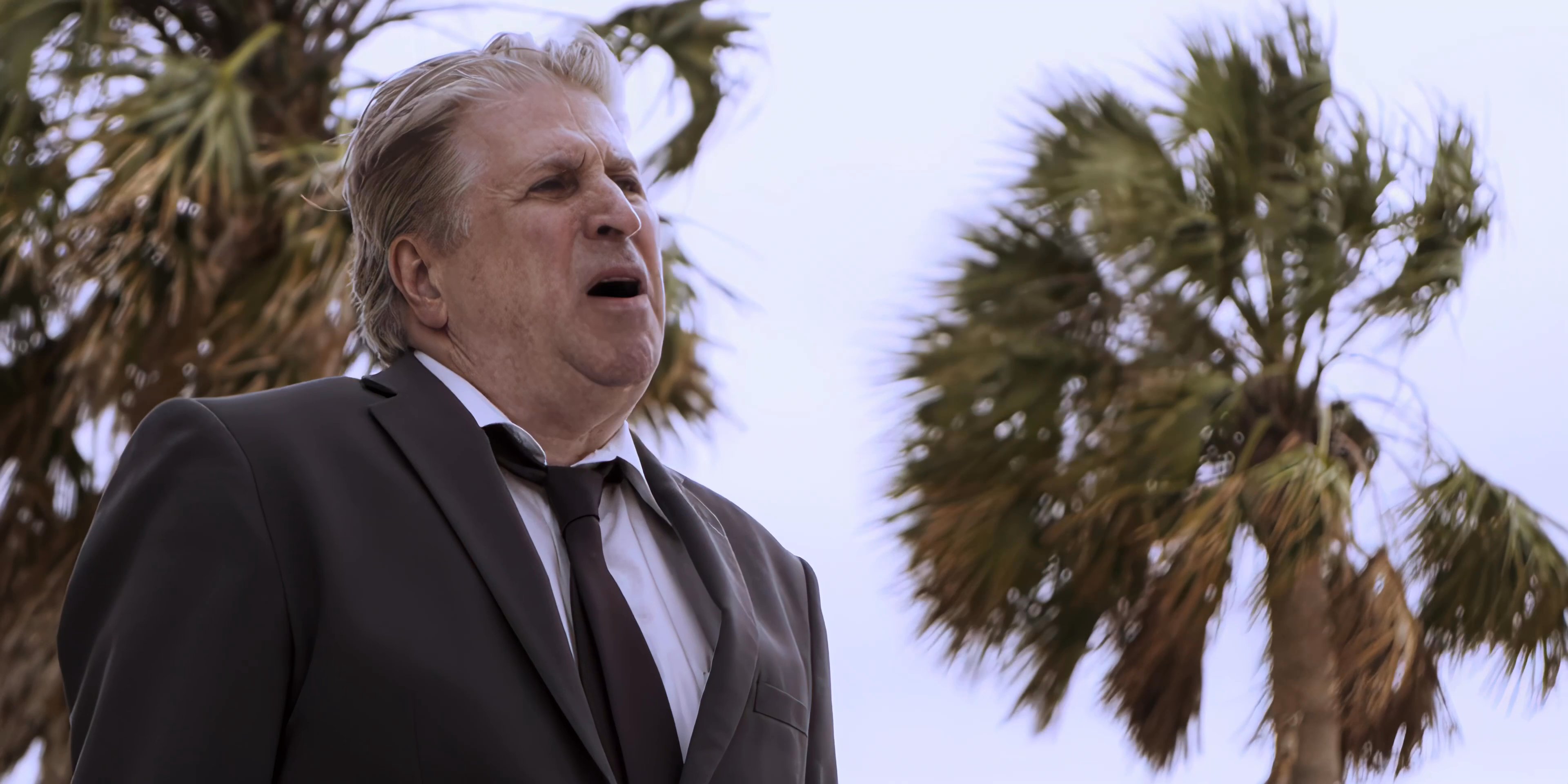 A man wearing a suit stands outdoors near palm trees, looking upward with a thoughtful and contemplative expression.