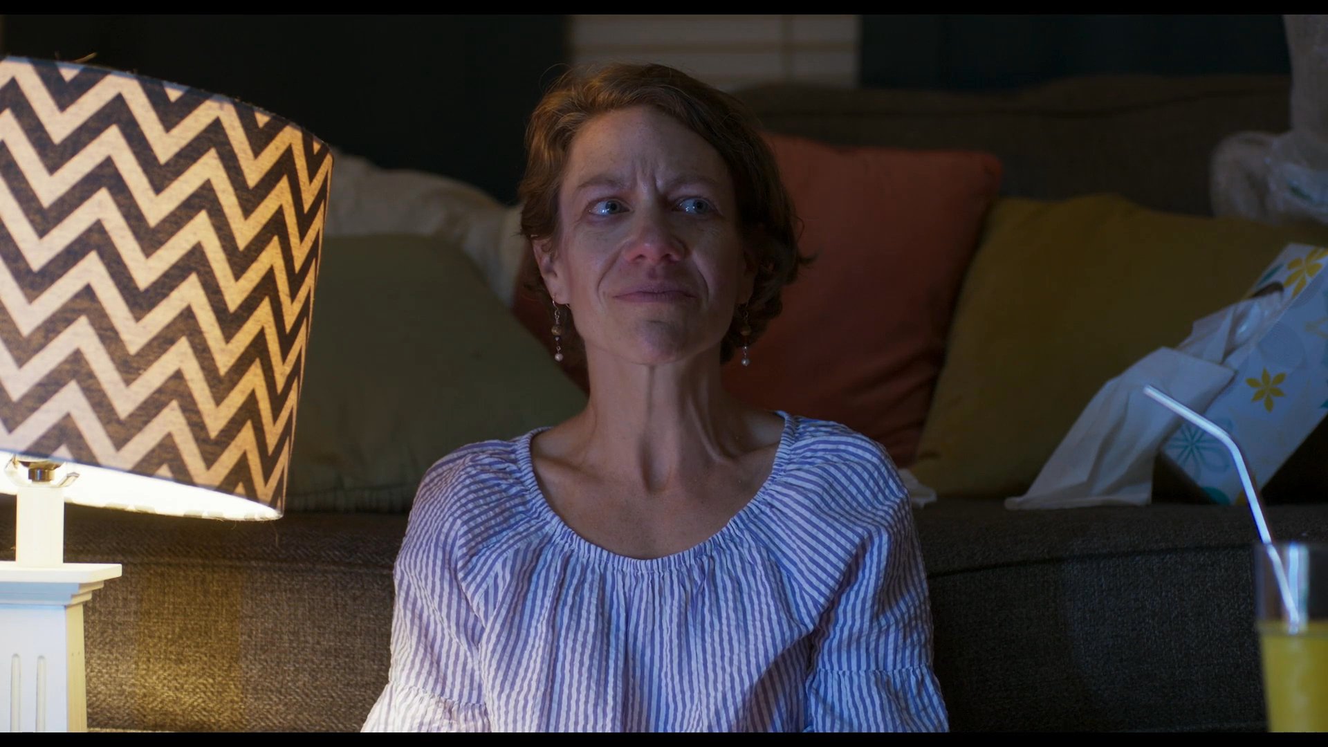 A woman sits on a sofa in a dimly lit living room, relaxing and observing her surroundings while holding a glass of juice.
