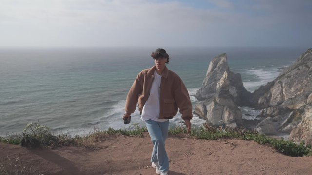 A curly-haired guy waves goodbye while walking on the edge of the cliff