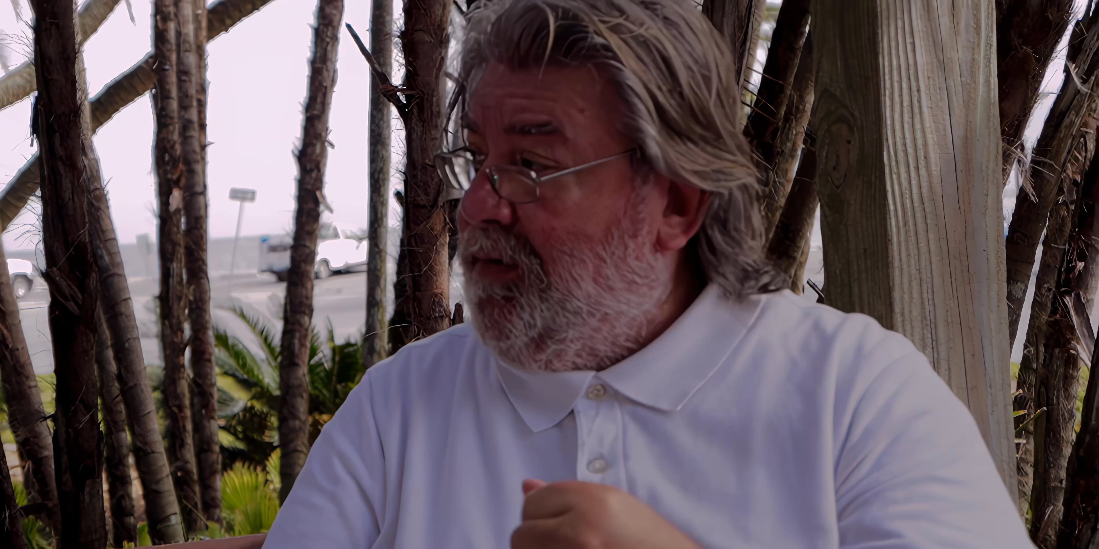 Two men engage in an earnest conversation while sitting outdoors near palm trees. They exchange thoughts with thoughtful gestures and focused expressions.