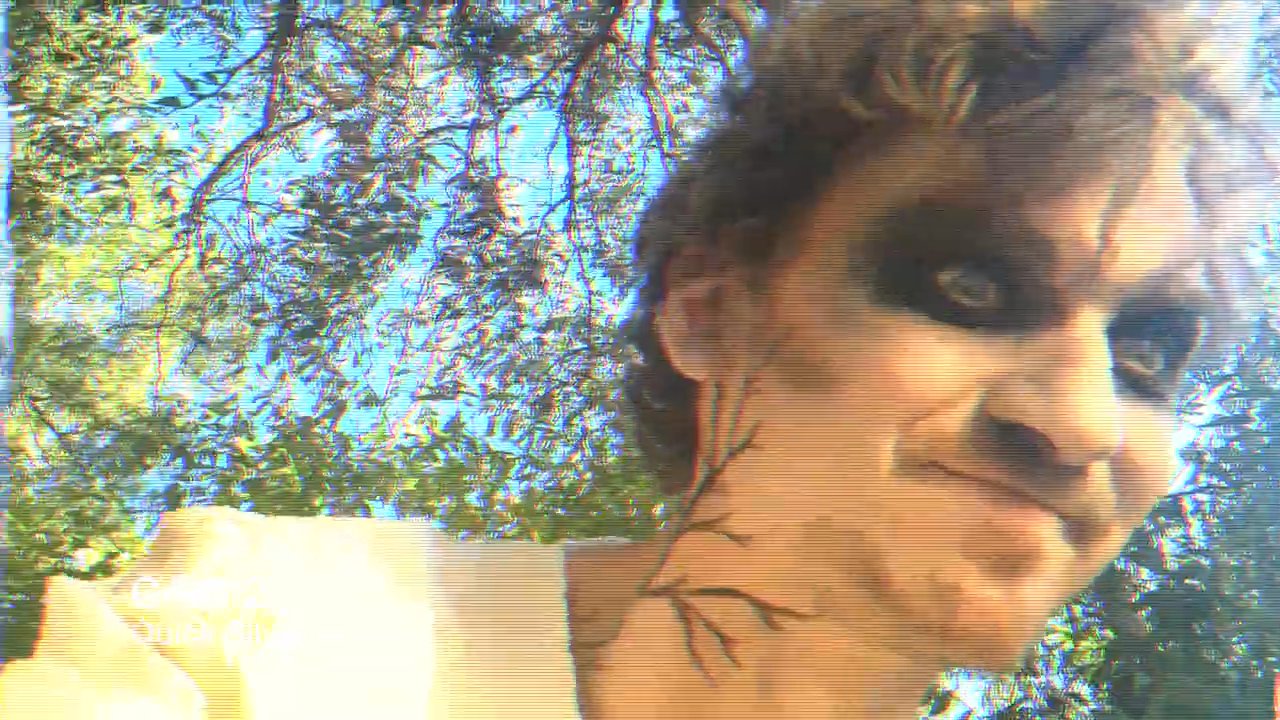 A performer with theatrical makeup interacts with the camera in a park environment while experiencing intermittent visual signal distortions. The sequence alternates between close-up character expressions and atmospheric outdoor shots.