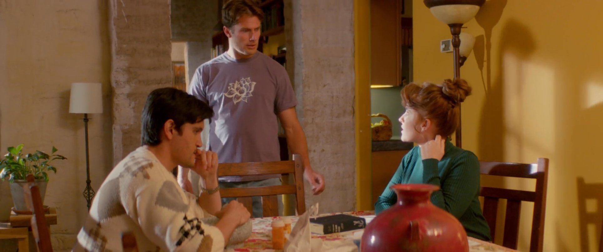Two individuals sit at a dining table while a third person initiates an exchange involving books within a home setting.