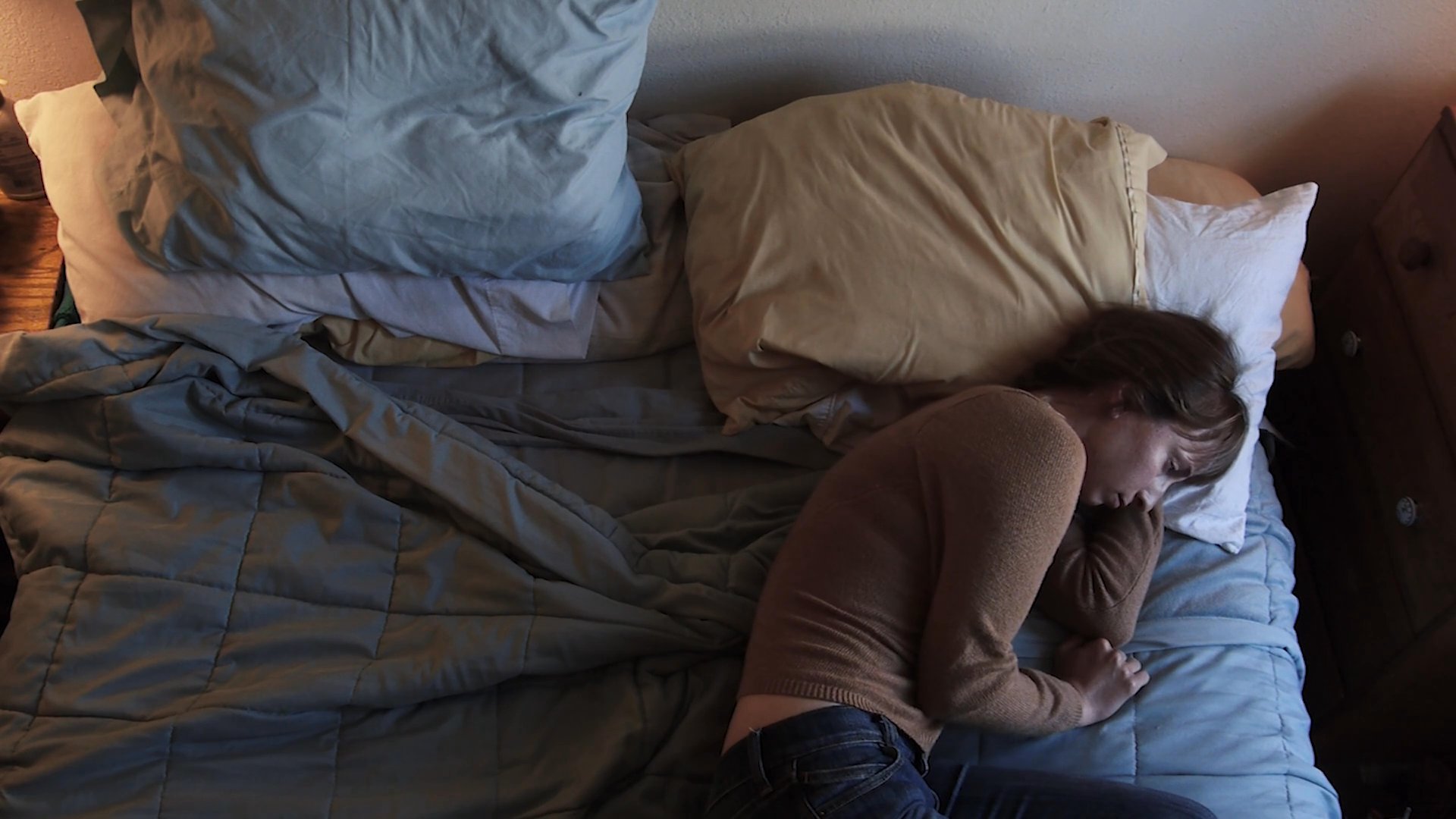 A woman struggles to rest in bed before transitioning to a somber, solitary afternoon at an outdoor park bench. The shift captures a sense of aimless isolation and quiet contemplation.