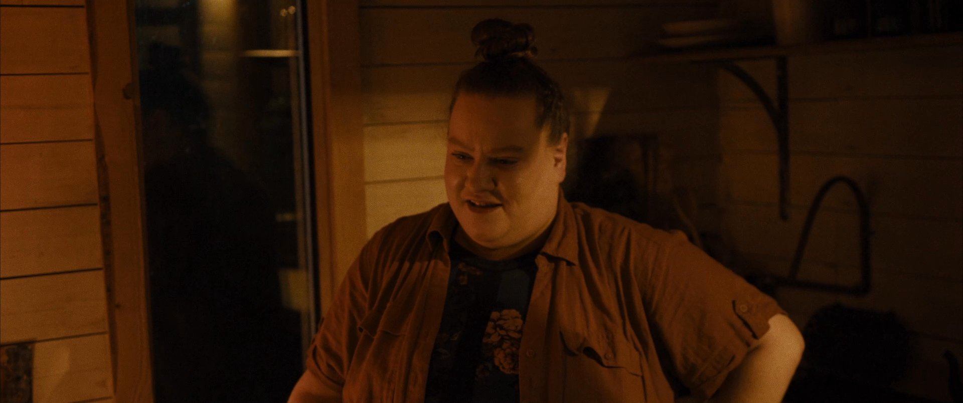 Two individuals engage in a quiet, intimate moment within a rustic, dimly lit cabin. They share a brief embrace before transitioning into a casual conversation.