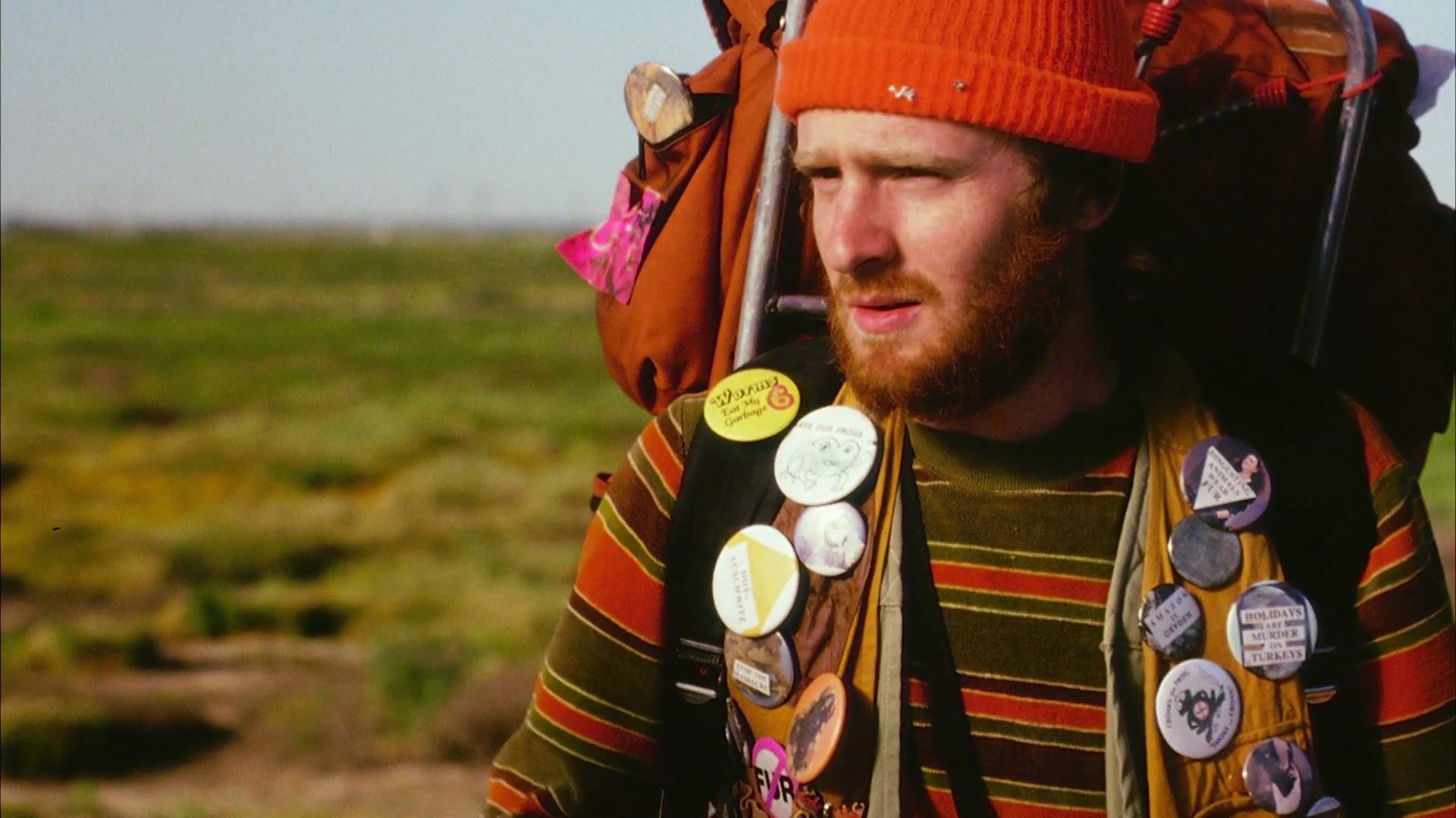 A cyclist carrying heavy gear stops on a remote road to interact with a pedestrian walking through a rural landscape. The two engage in a brief, informal conversation under the open sky.