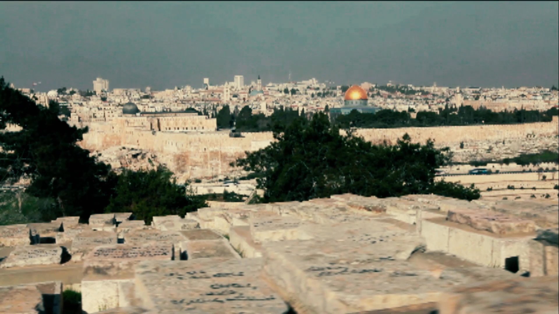 An interviewer engages in a structured discussion regarding historical events, transitioning between an indoor setting and outdoor location footage of Jerusalem and ancient stone architecture.