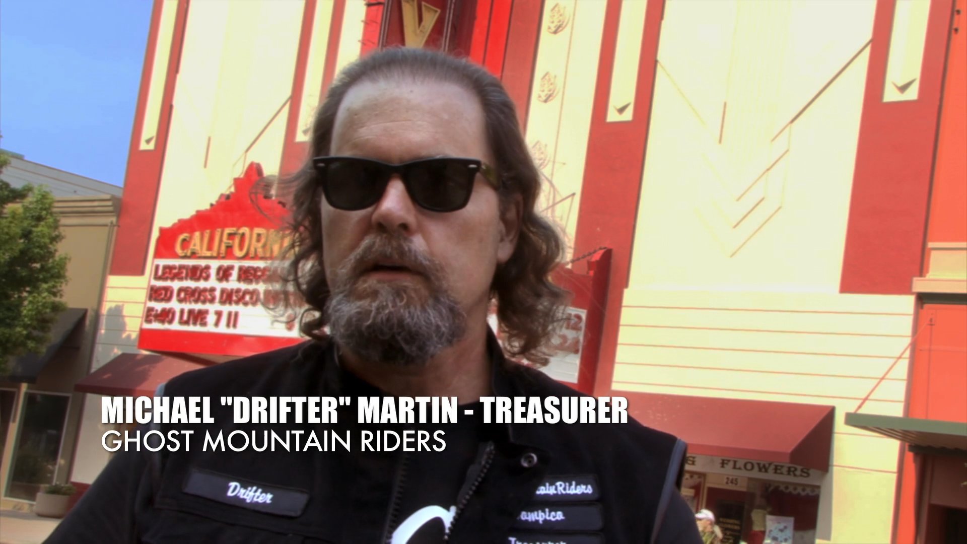Law enforcement officers gather outside a public venue while Michael 'Drifter' Martin, a representative of the Ghost Mountain Riders, provides an interview regarding the event.