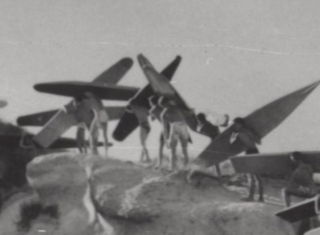 Athletes carry surfboards along a coastal cliff in vintage black-and-white film, followed by an elderly man reflecting on the experience during an interview.