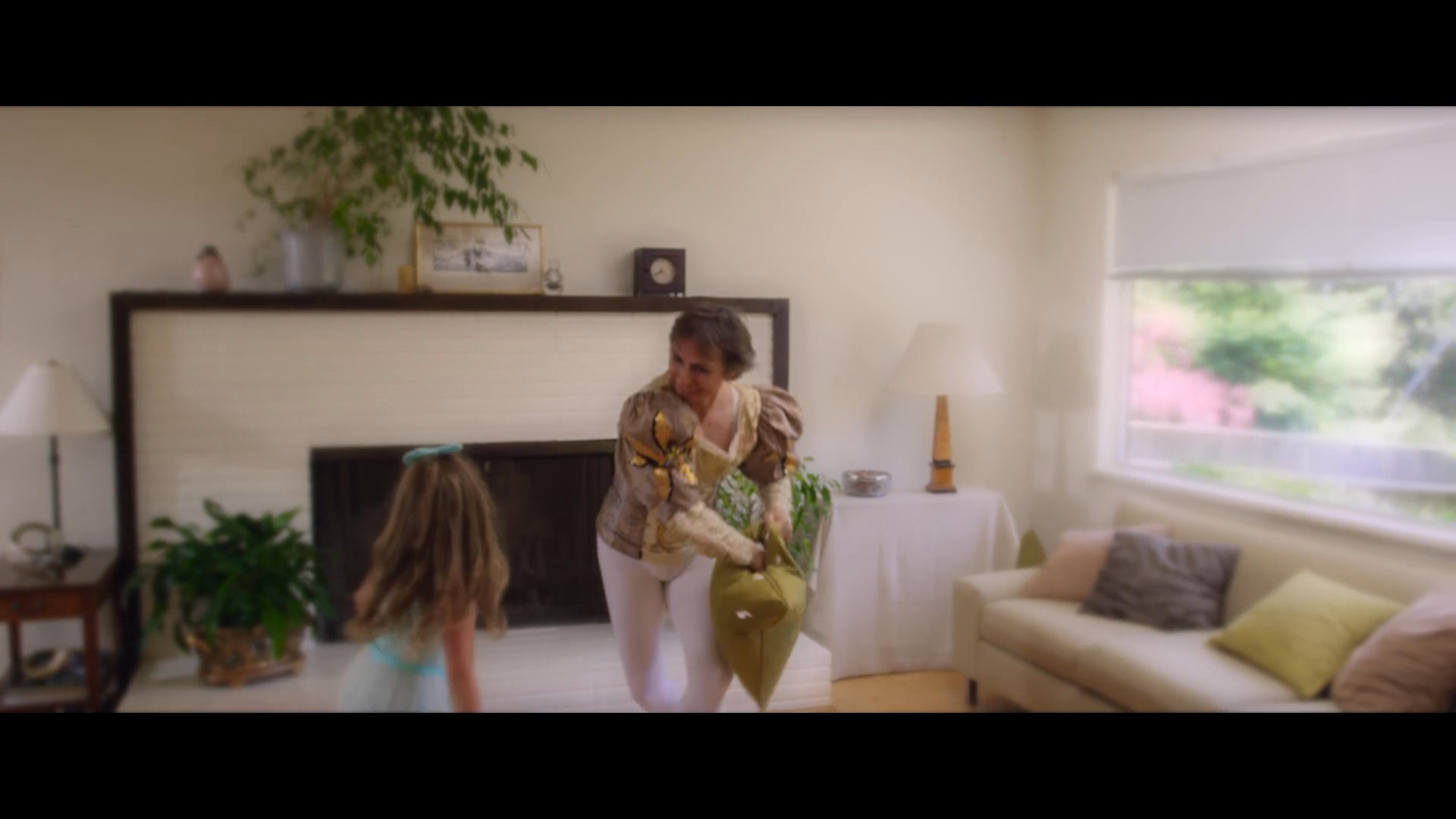 Two children engage in a playful pillow fight in a sunlit living room, scattering feathers throughout the space while jumping on furniture.