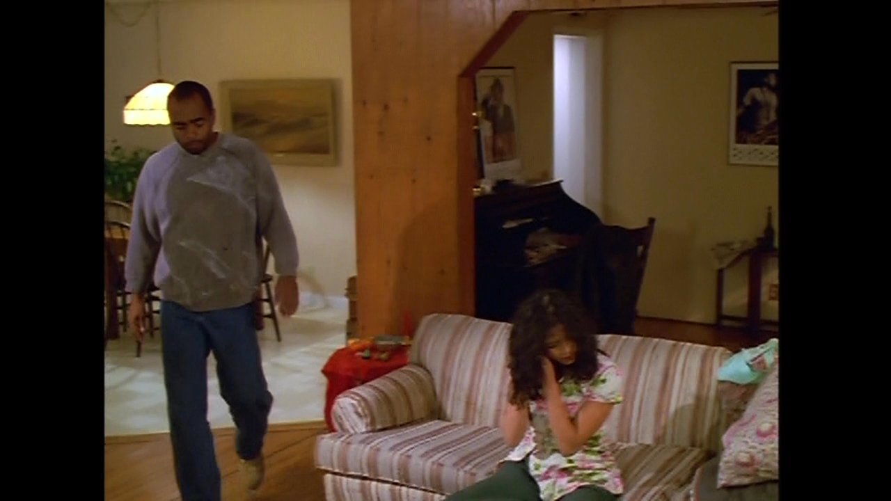 A man speaks intensely on a telephone while a woman sits distressed on a sofa in a living room. The man then approaches her, creating a moment of palpable interpersonal tension.