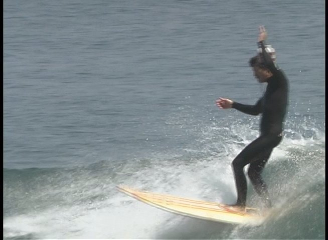 A surfer maneuvers a longboard across ocean waves while wearing a wetsuit, interspersed with an interview segment featuring a man wearing sunglasses.