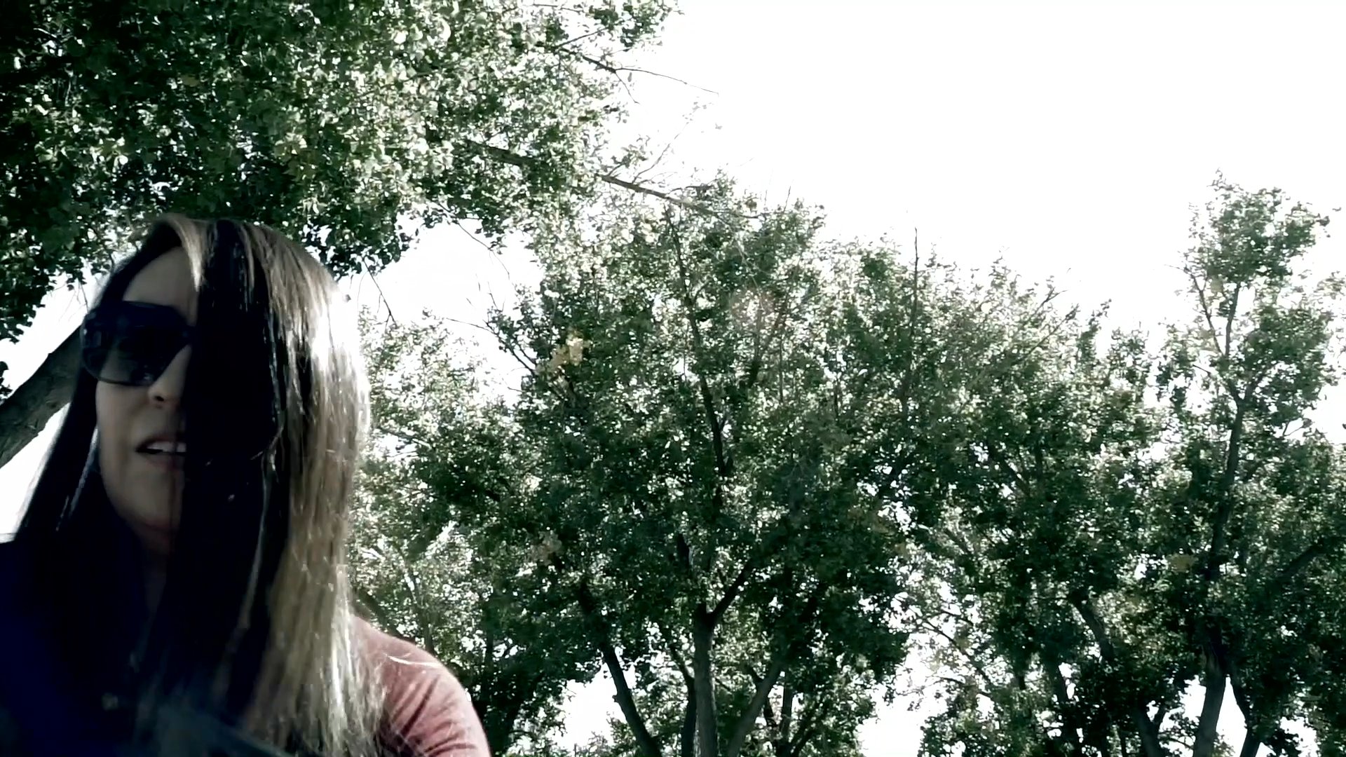 A woman wearing sunglasses walks through a sun-dappled yard filled with mature trees. The camera moves through the natural landscape, capturing the bright daylight and shaded areas.