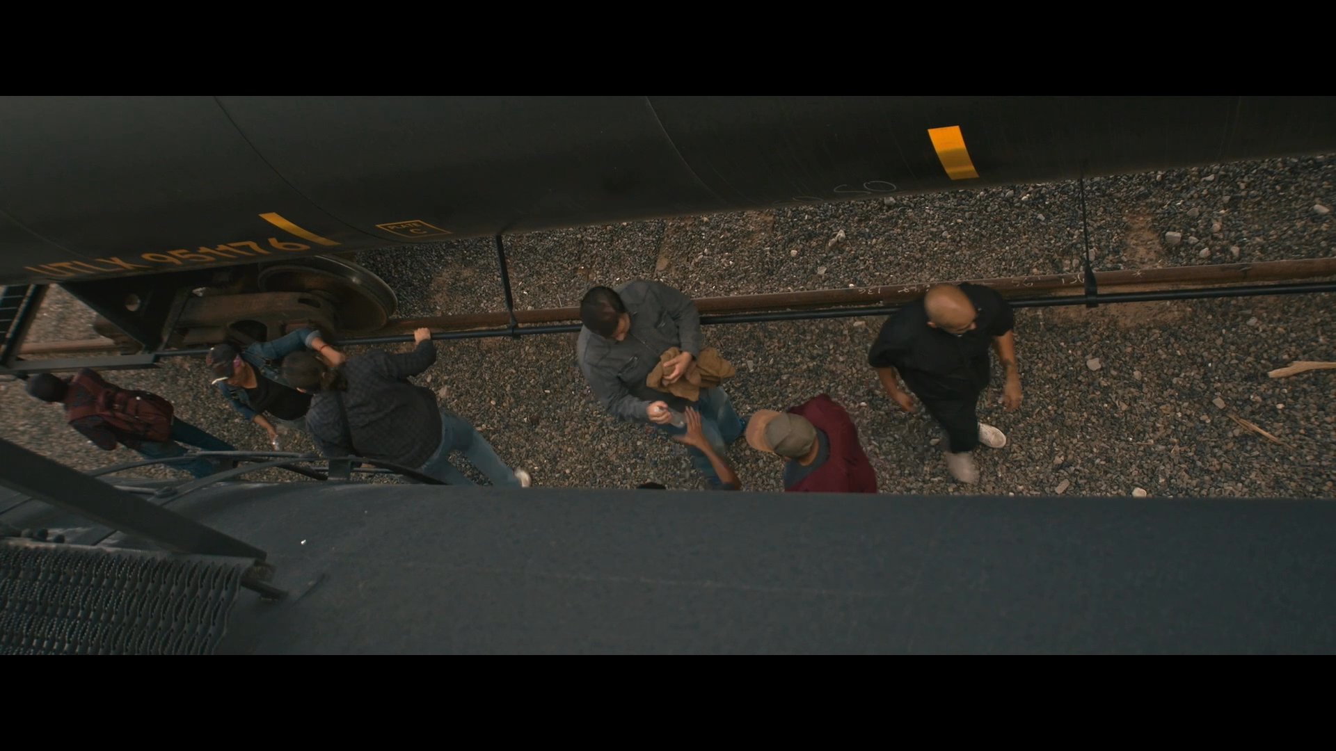 Volunteers welcome arriving migrants at a railroad tracks, offering food and water to those in need. The scene focuses on the supportive reception and the ongoing conversation between the groups.