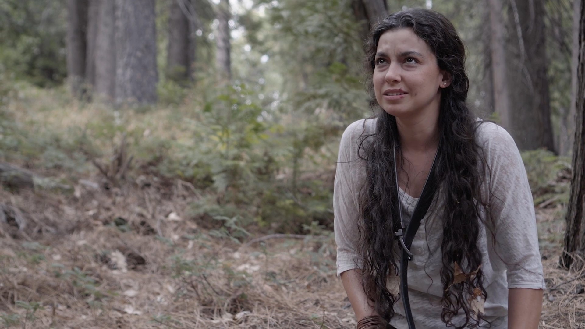 A distraught woman sits alone in a wooded area, experiencing intense emotional distress and sobbing. Her body language reflects profound sorrow as she looks around the forest environment.
