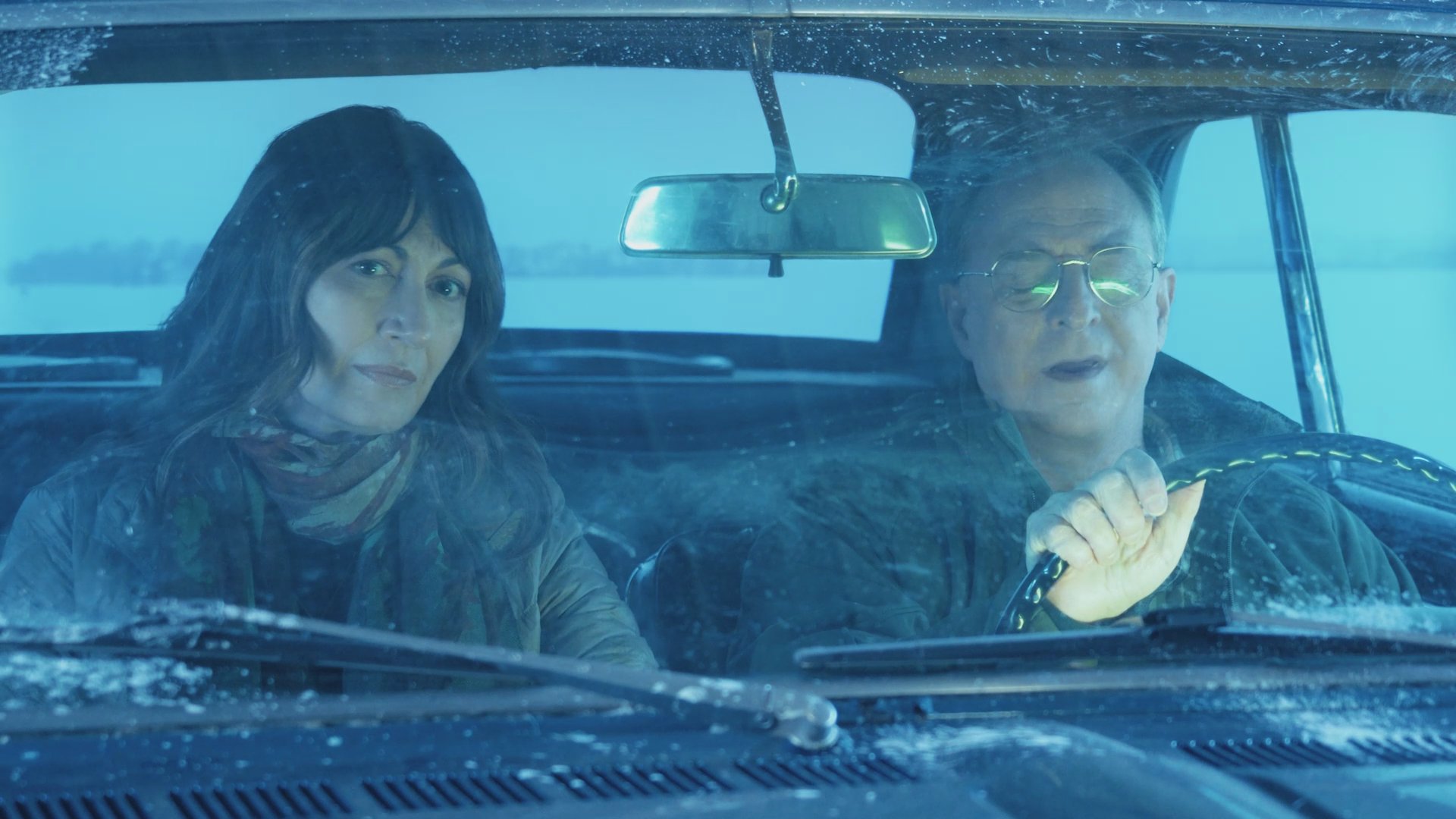 A man and woman travel in a classic car along a desolate, snow-covered highway. The pair engages in conversation while navigating the wintry conditions.