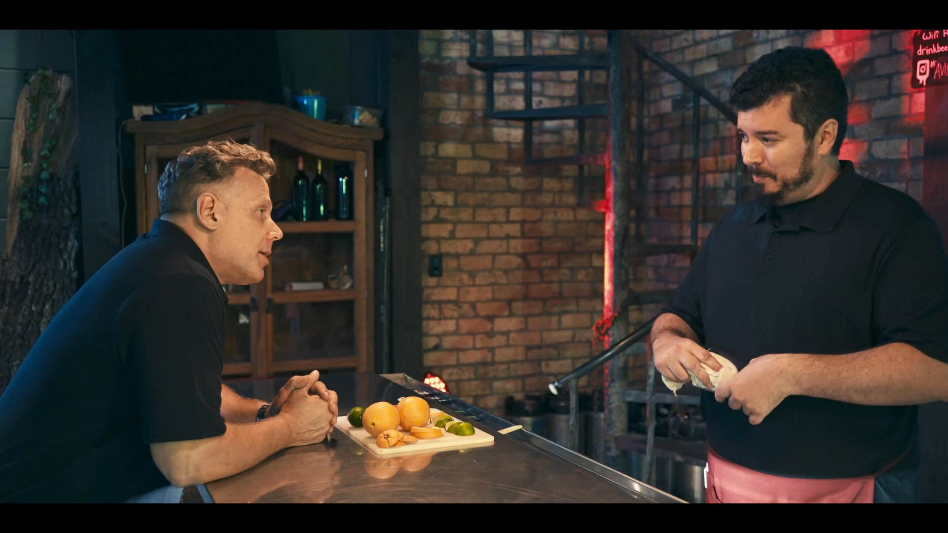 A bartender converses with a customer sitting at a bar while preparing a beverage, maintaining a professional and engaging service environment.