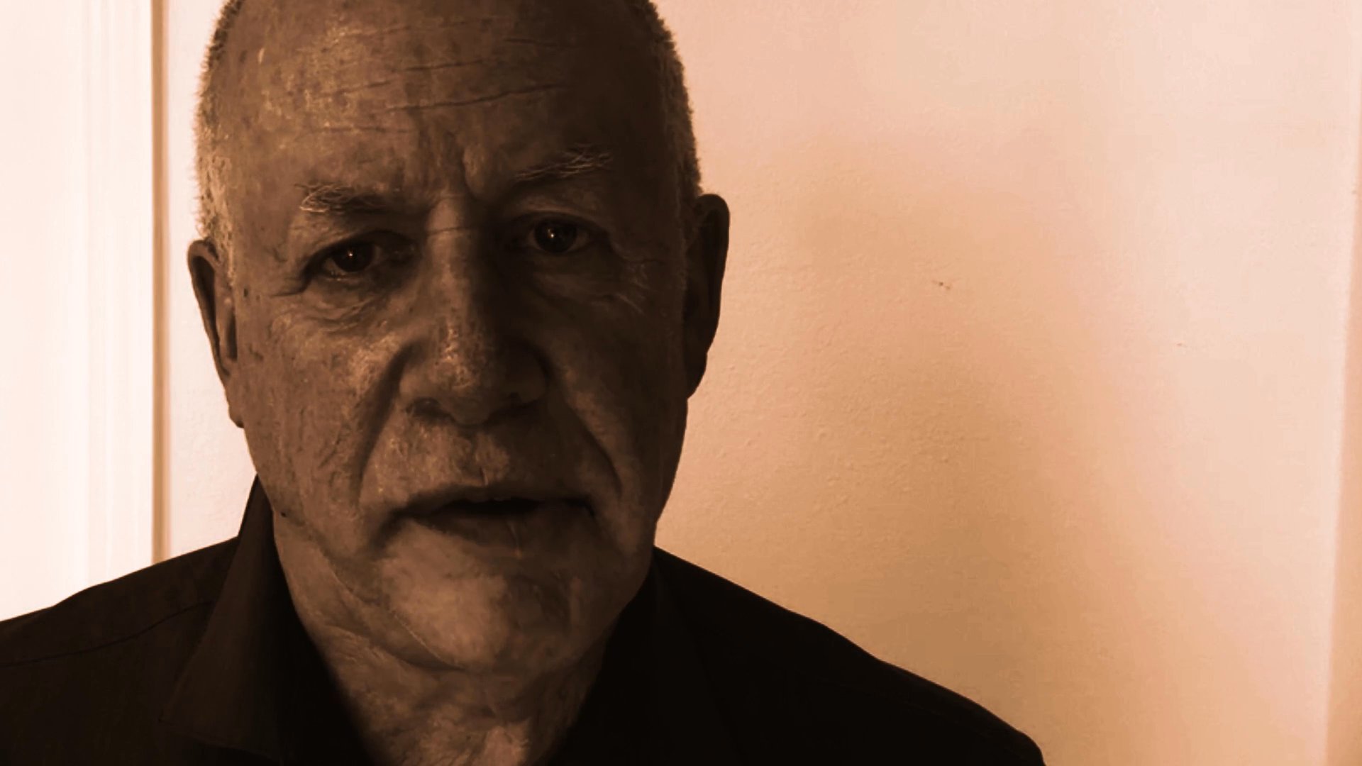An older man faces the camera while speaking directly to the audience in a sepia-toned, close-up shot.