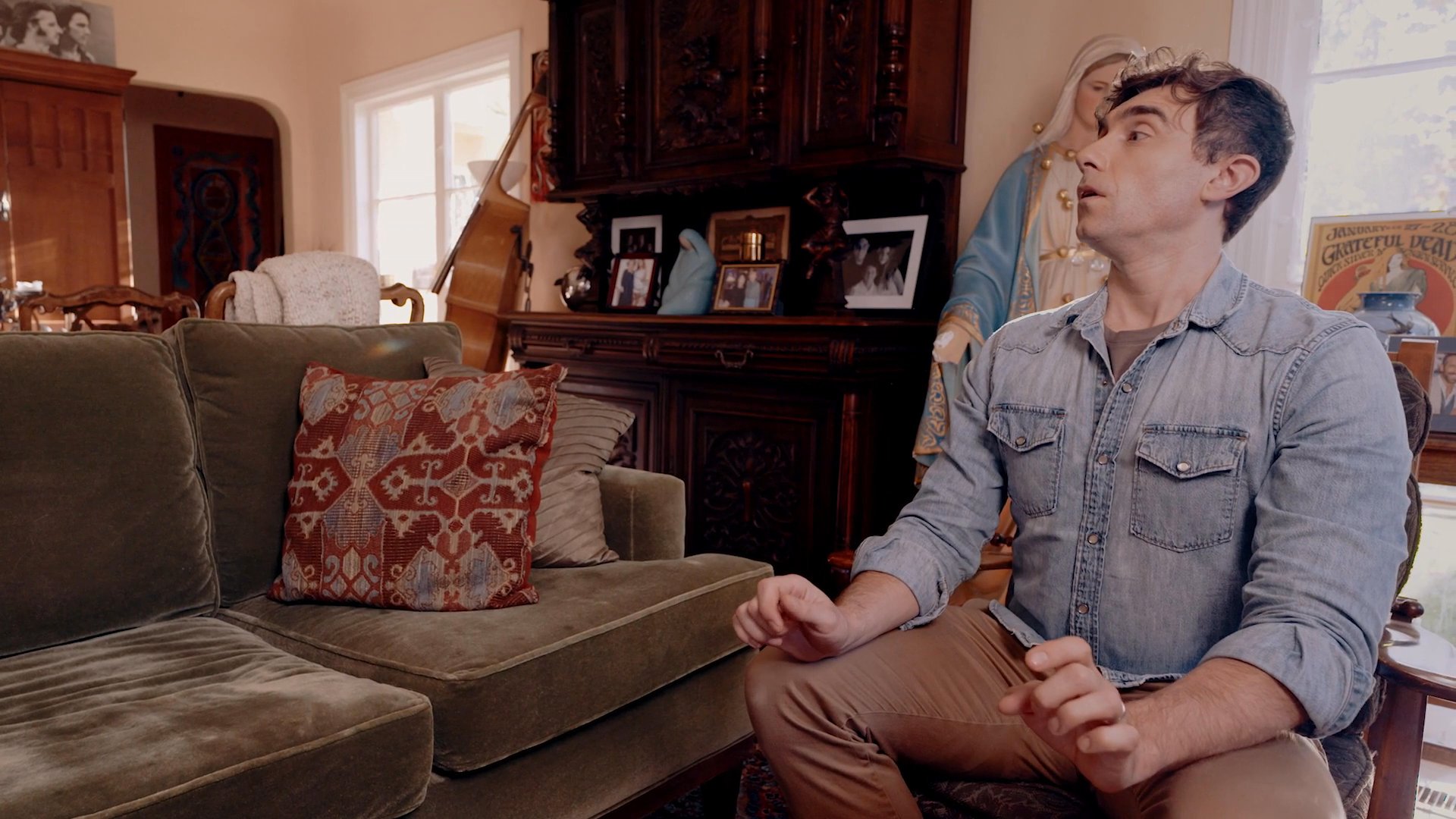 A man sits on a couch in a living room, gesturing while speaking, before transitioning to an office setting where he approaches a reception desk.