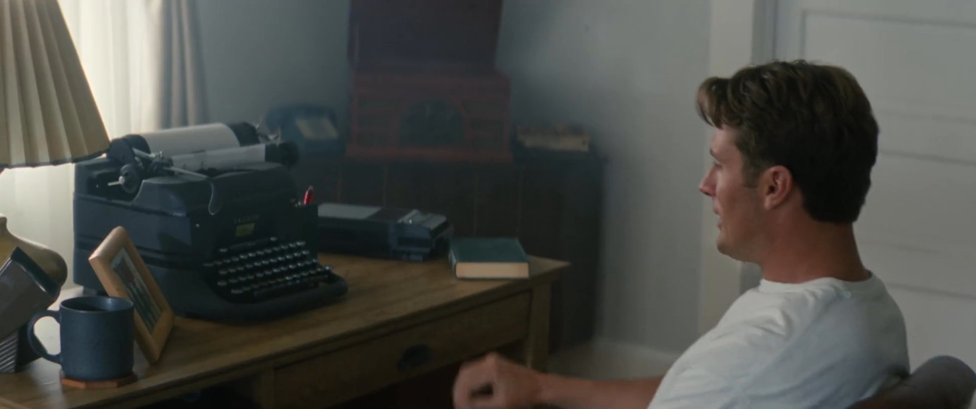A man pauses from writing at a vintage typewriter to contemplate a framed photograph of a surfing group. He exits his coastal home, stepping outside to observe the ocean environment.